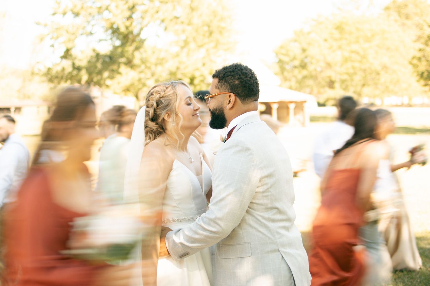 Bride and groom share a romantic moment surrounded by blurred wedding guests outdoors in golden sunlight.