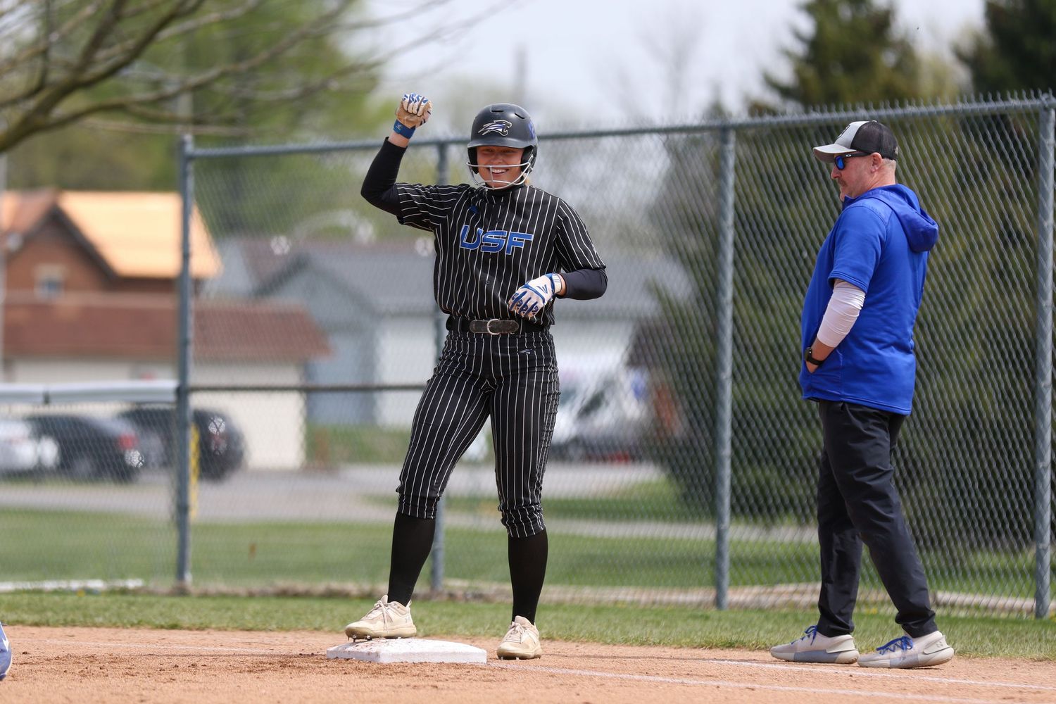 Baseball player in pinstriped uniform stands at base with raised arm as coach looks on during game.