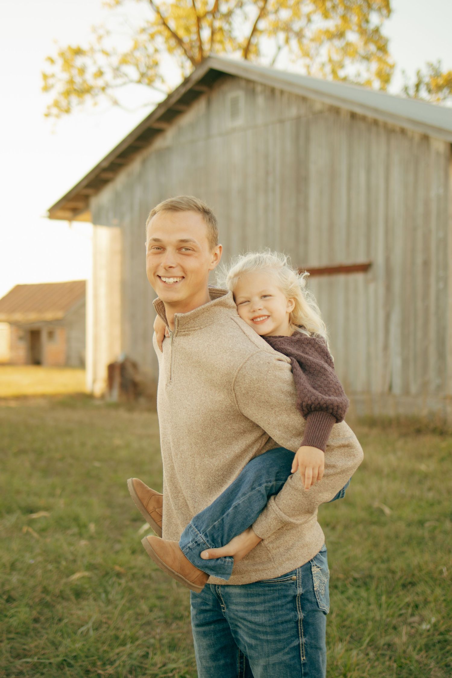 Smiling man holding young blonde girl on a farm with rustic barn in background during golden hour.