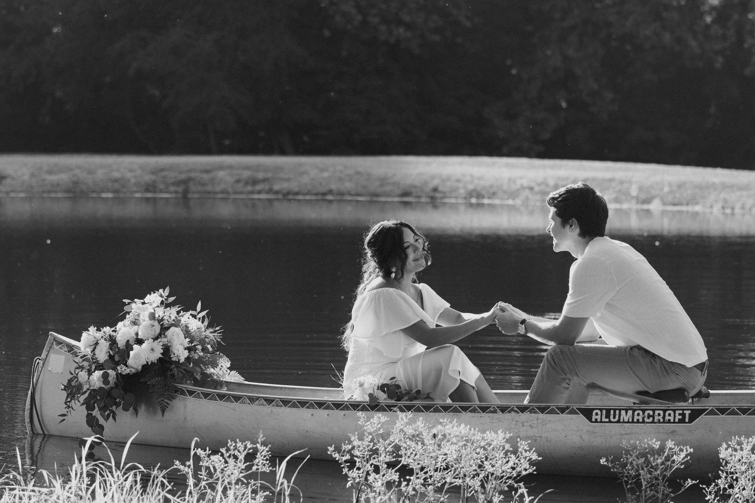 Black and white photo of a couple holding hands in a canoe on a lake with a floral bouquet nearby.