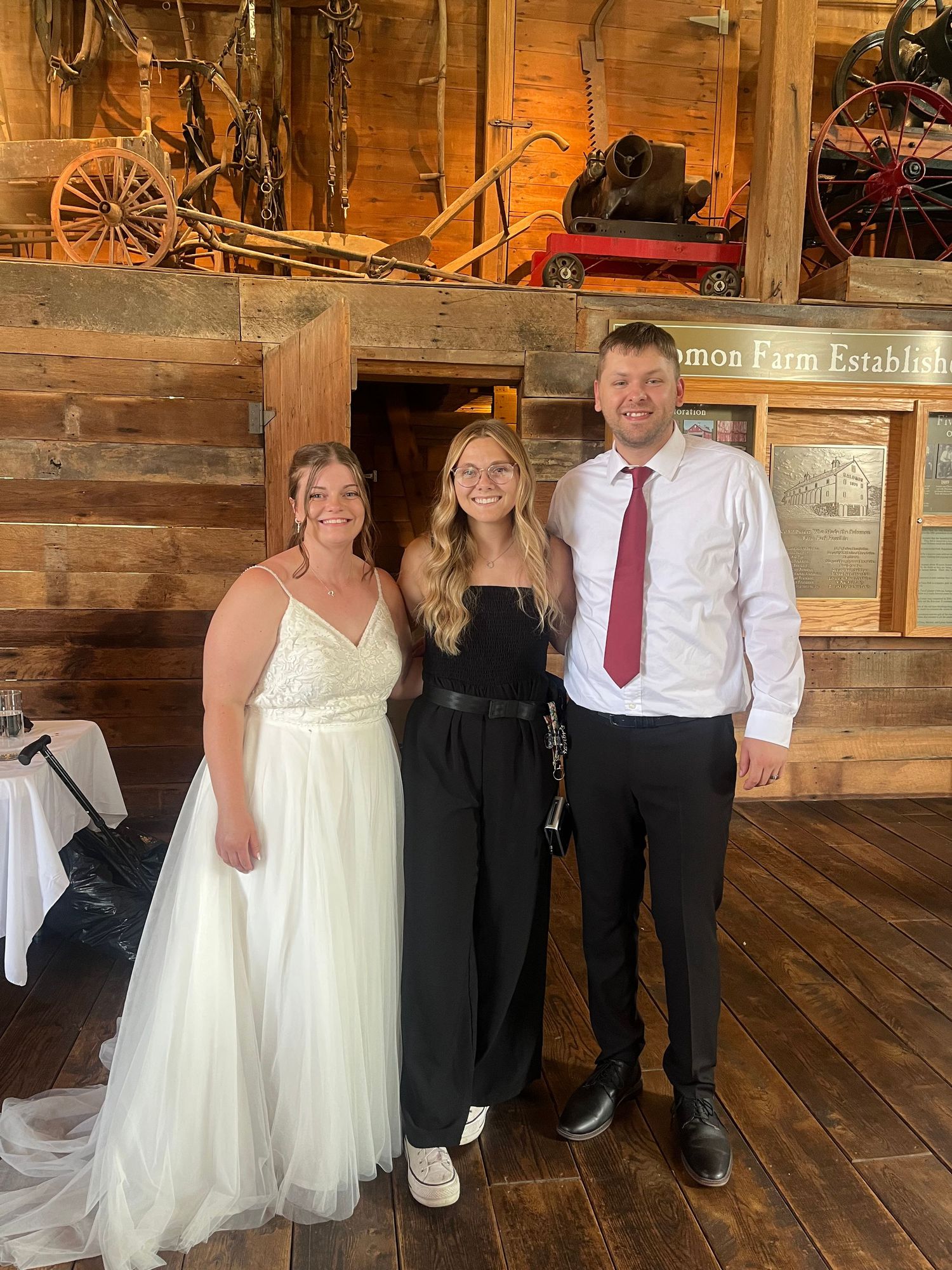 Bride in white gown poses with two guests in rustic barn wedding venue with wooden walls and antique decor.