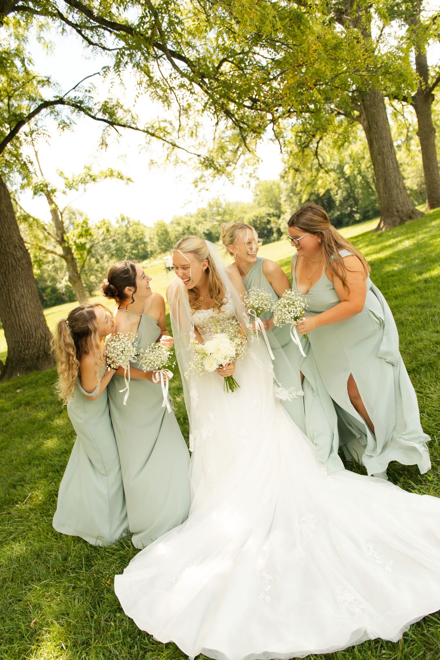 Bride in white gown with bridesmaids in sage green dresses holding white bouquets laughing outdoors under trees.