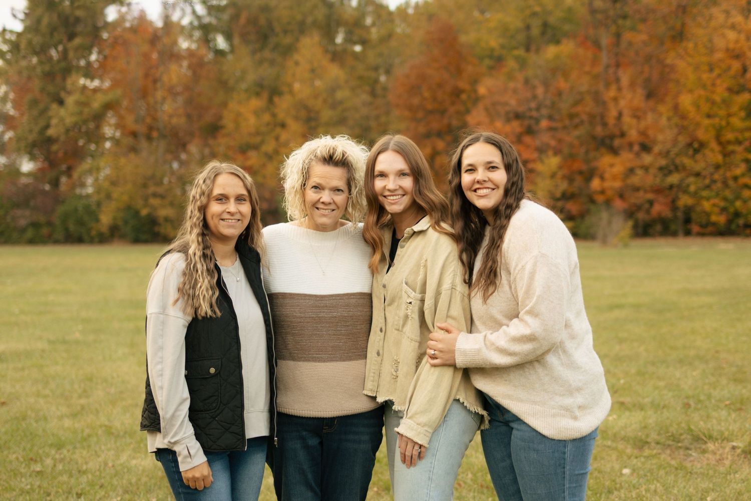 Four women smiling together outdoors in front of colorful autumn foliage on a green lawn.