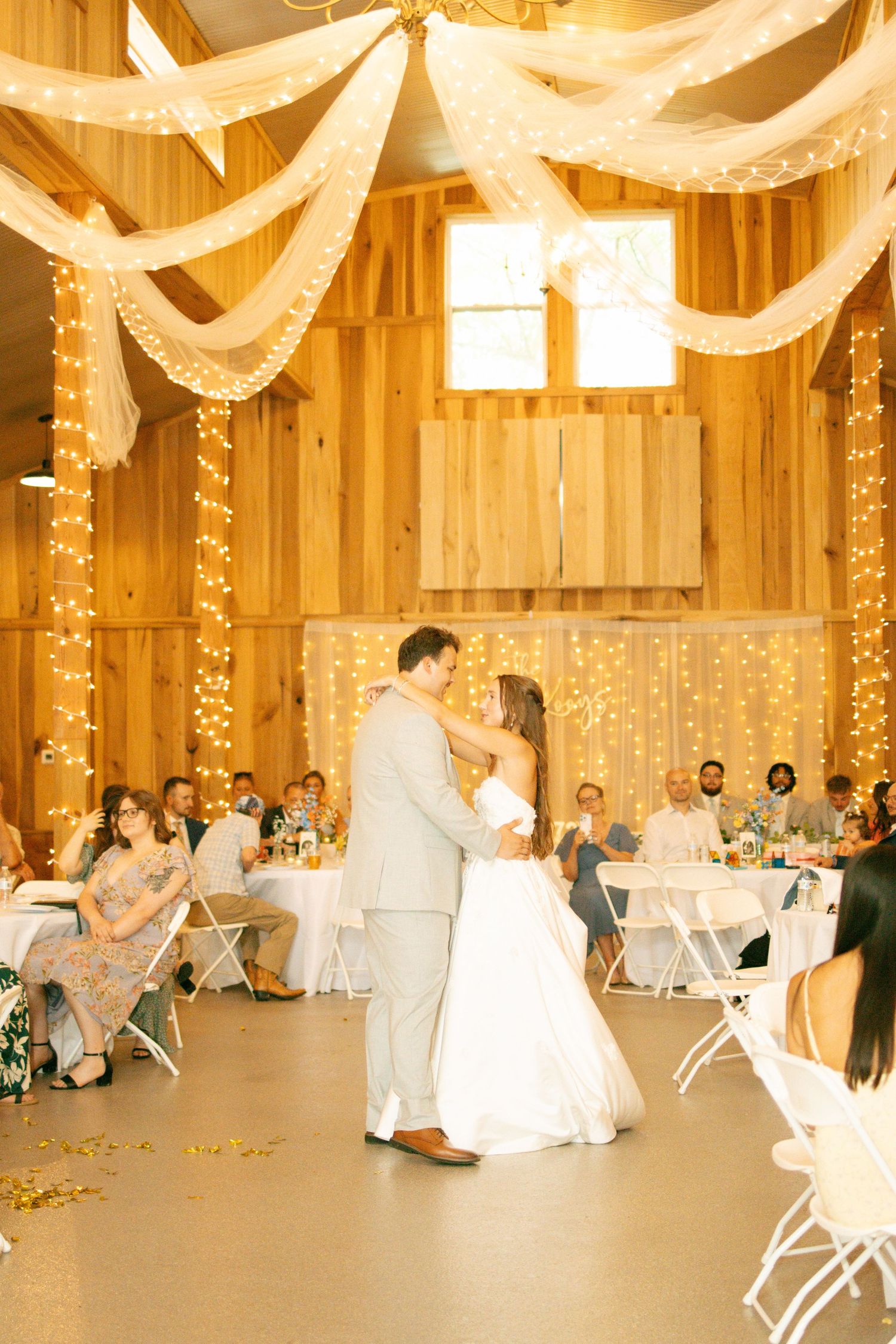 Bride and groom share first dance in a warmly lit wooden barn decorated with string lights and white draping.