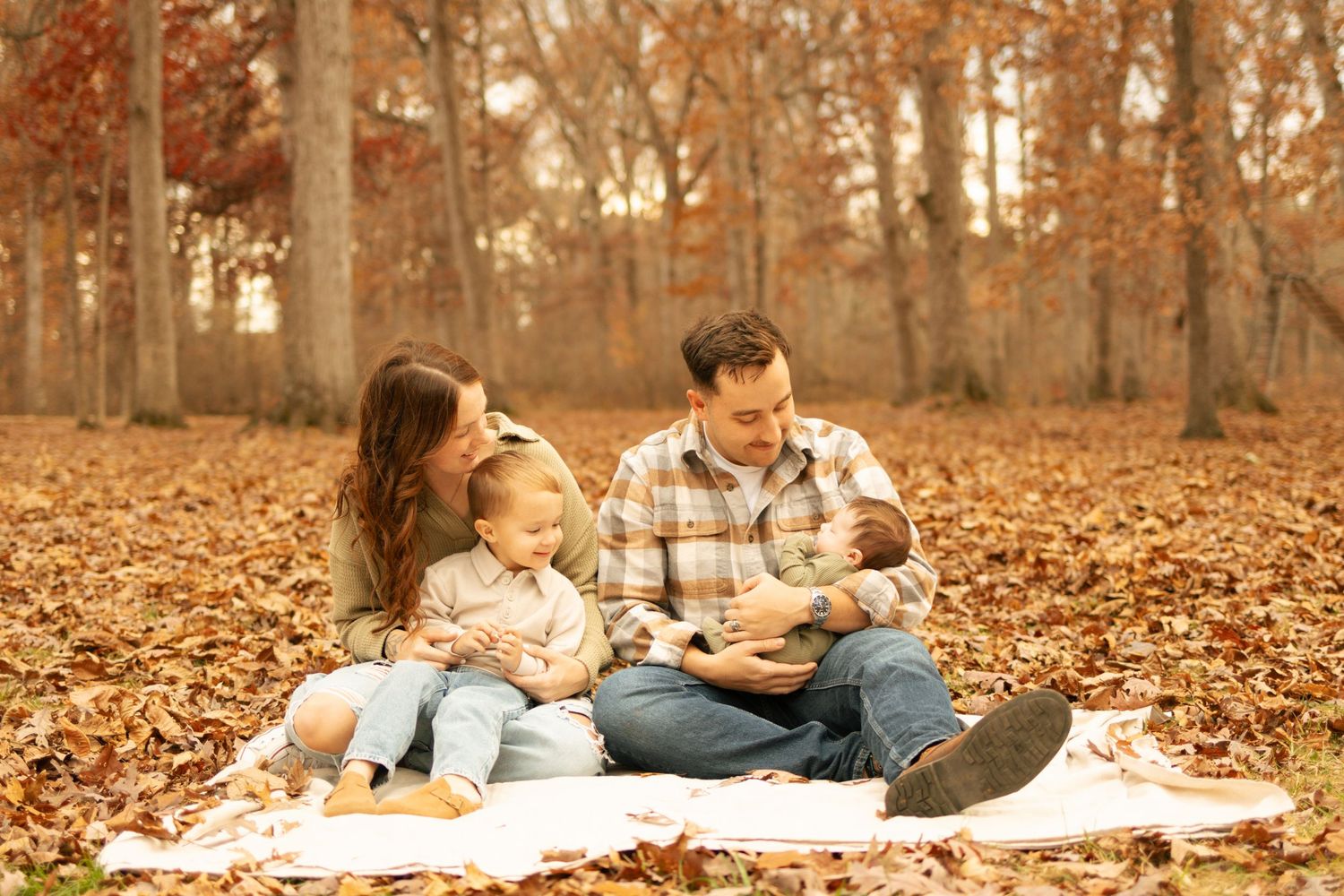 Family of four sitting on a blanket in autumn leaves, parents smiling at newborn baby with toddler beside them.