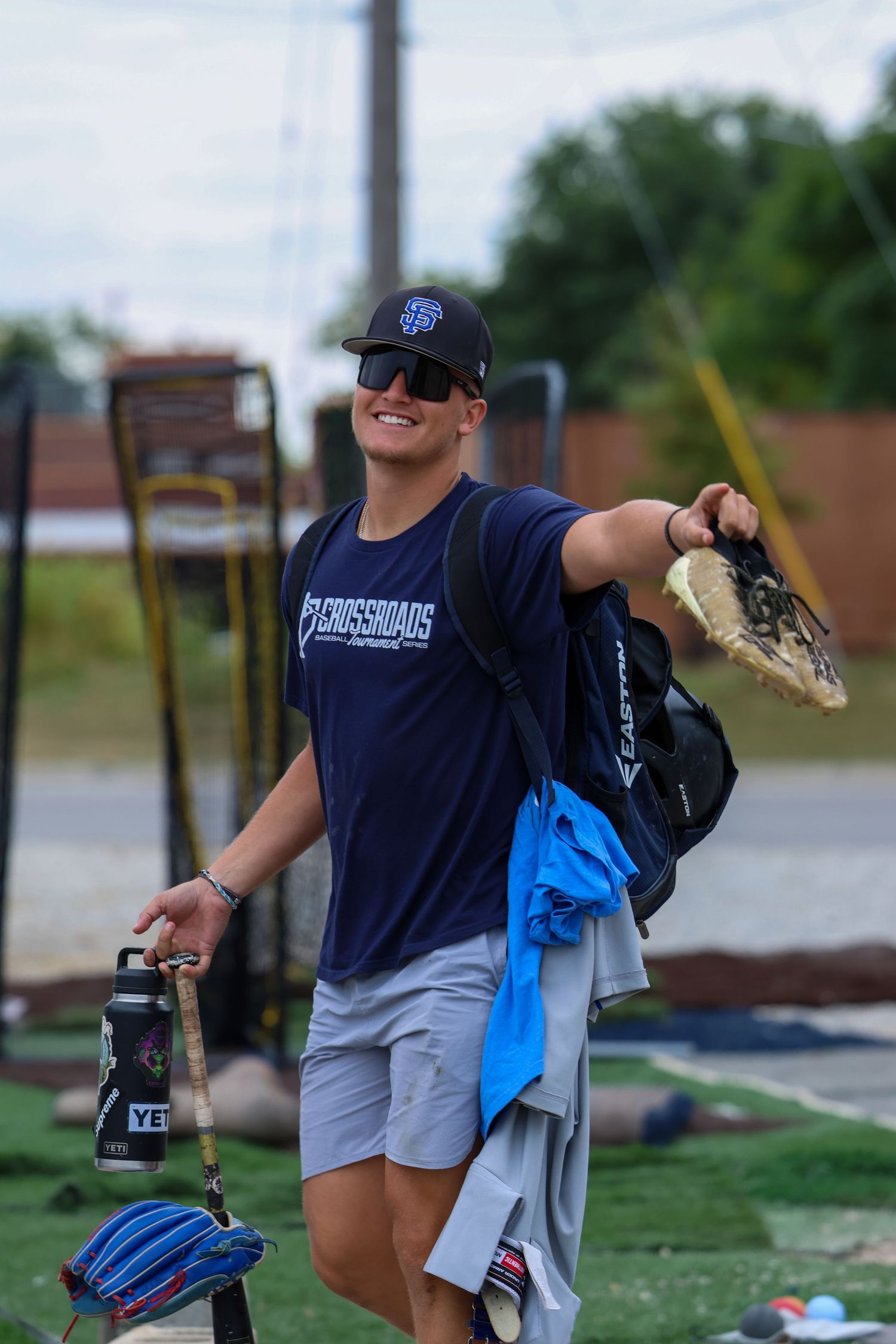 A baseball player in a navy t-shirt and grey shorts carries gear including a glove and backpack on a sports field.