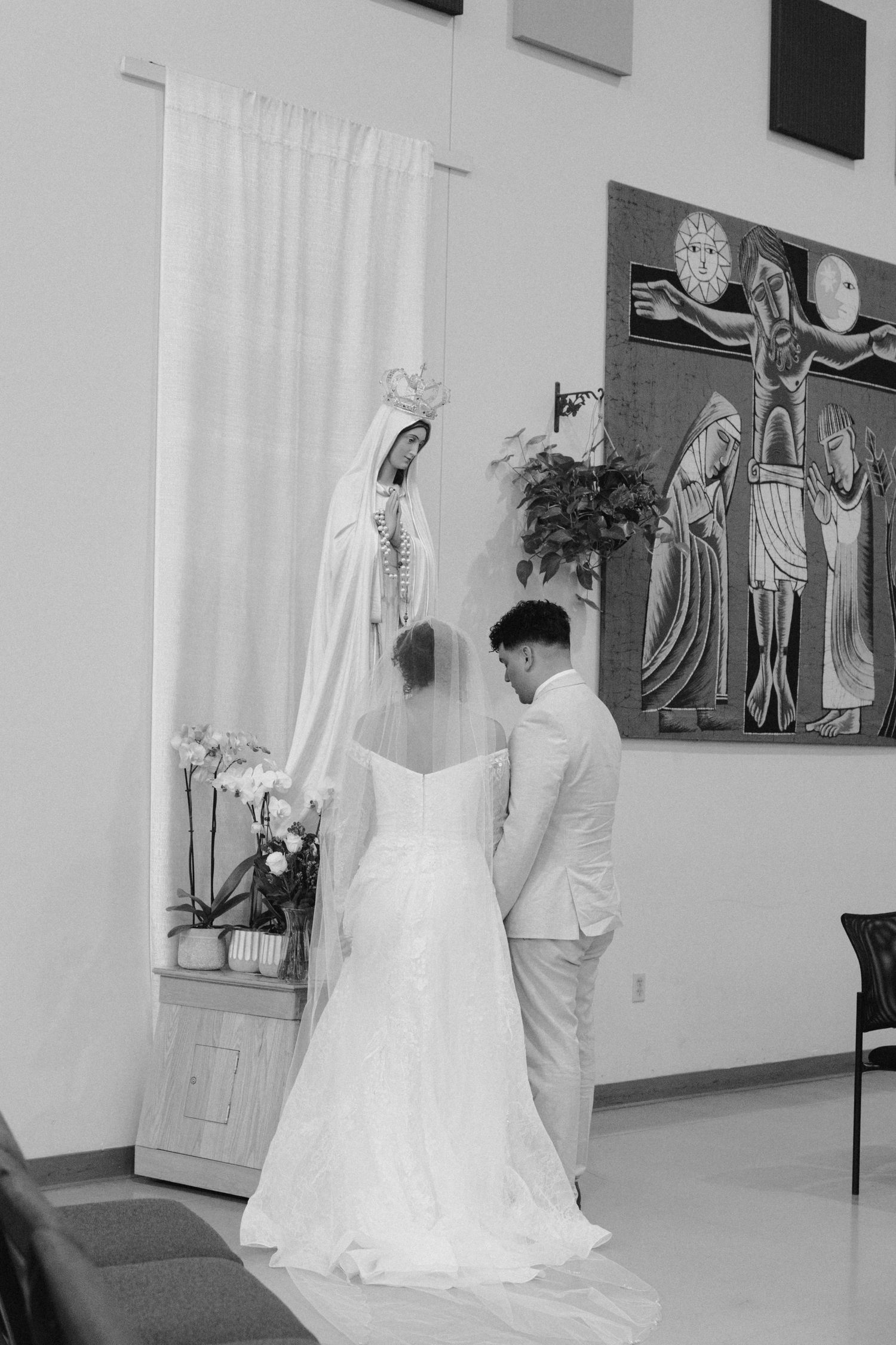 A bride and groom pray together in a church sanctuary before a statue of Mary and religious artwork.