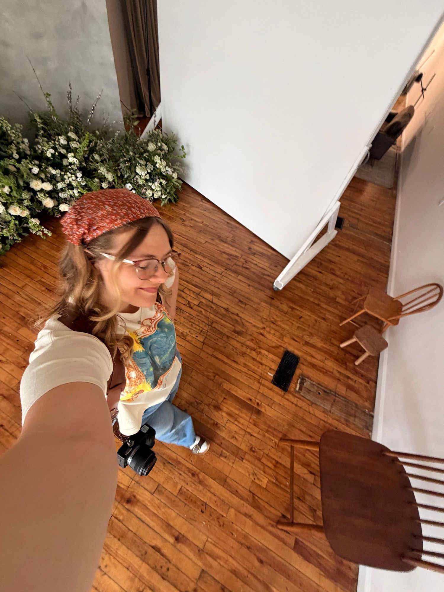Woman in glasses and red headband takes a selfie from above in a studio with hardwood floors and white backdrop.