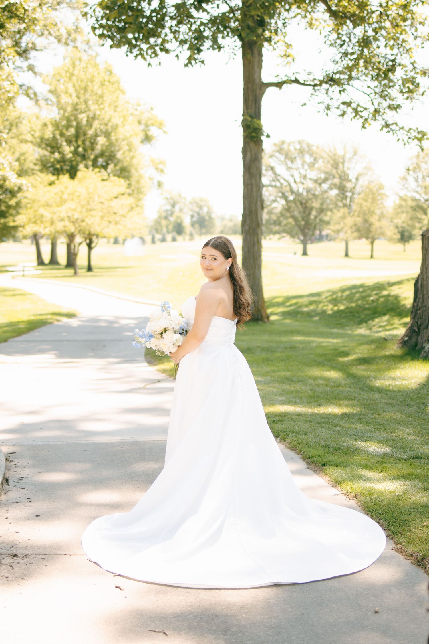 Bride in strapless white ball gown with long train holds bouquet on sunlit path surrounded by lush green trees.