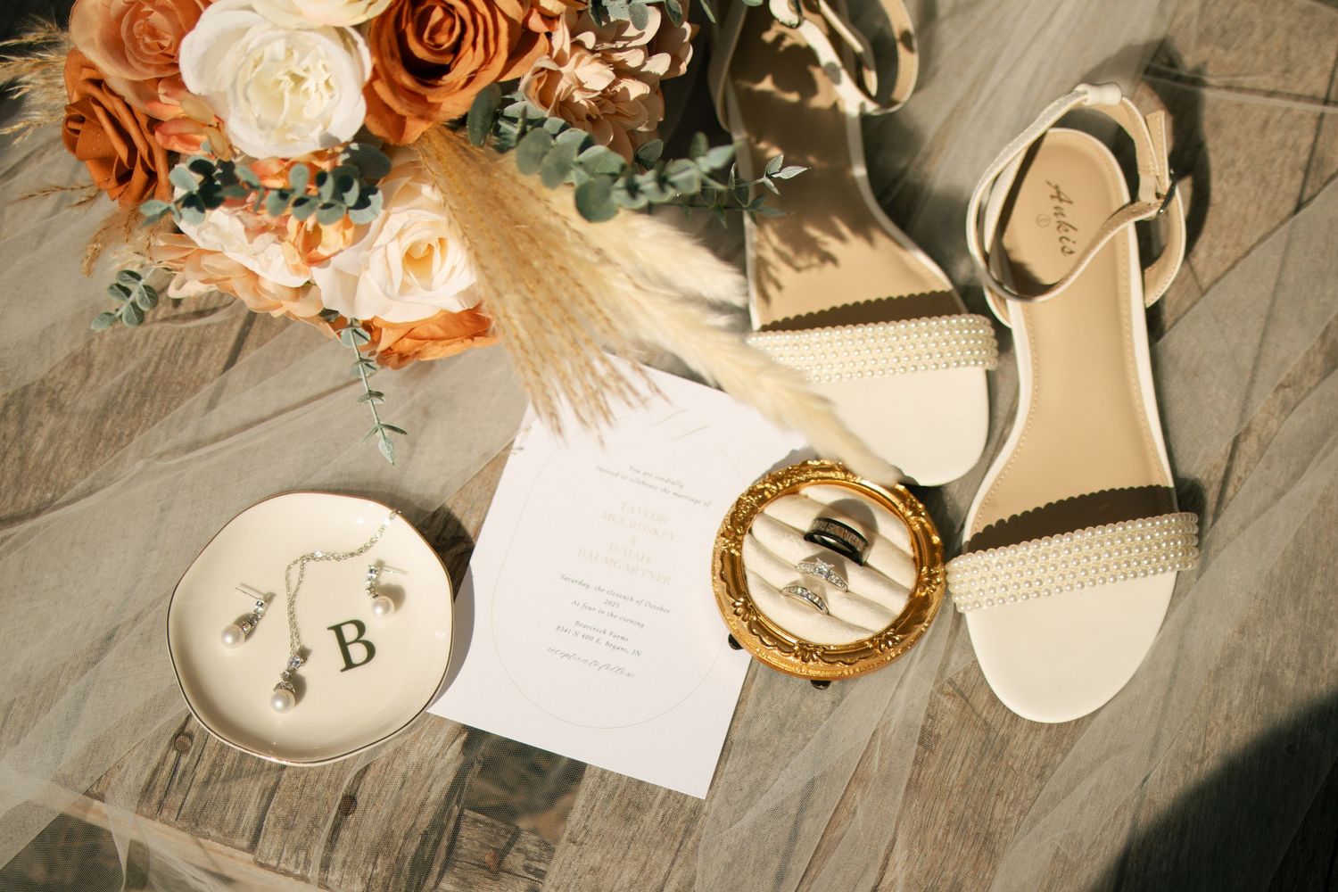 Elegant wedding flat lay with white heels, rings, floral bouquet, earrings, and invitation on wood floor.