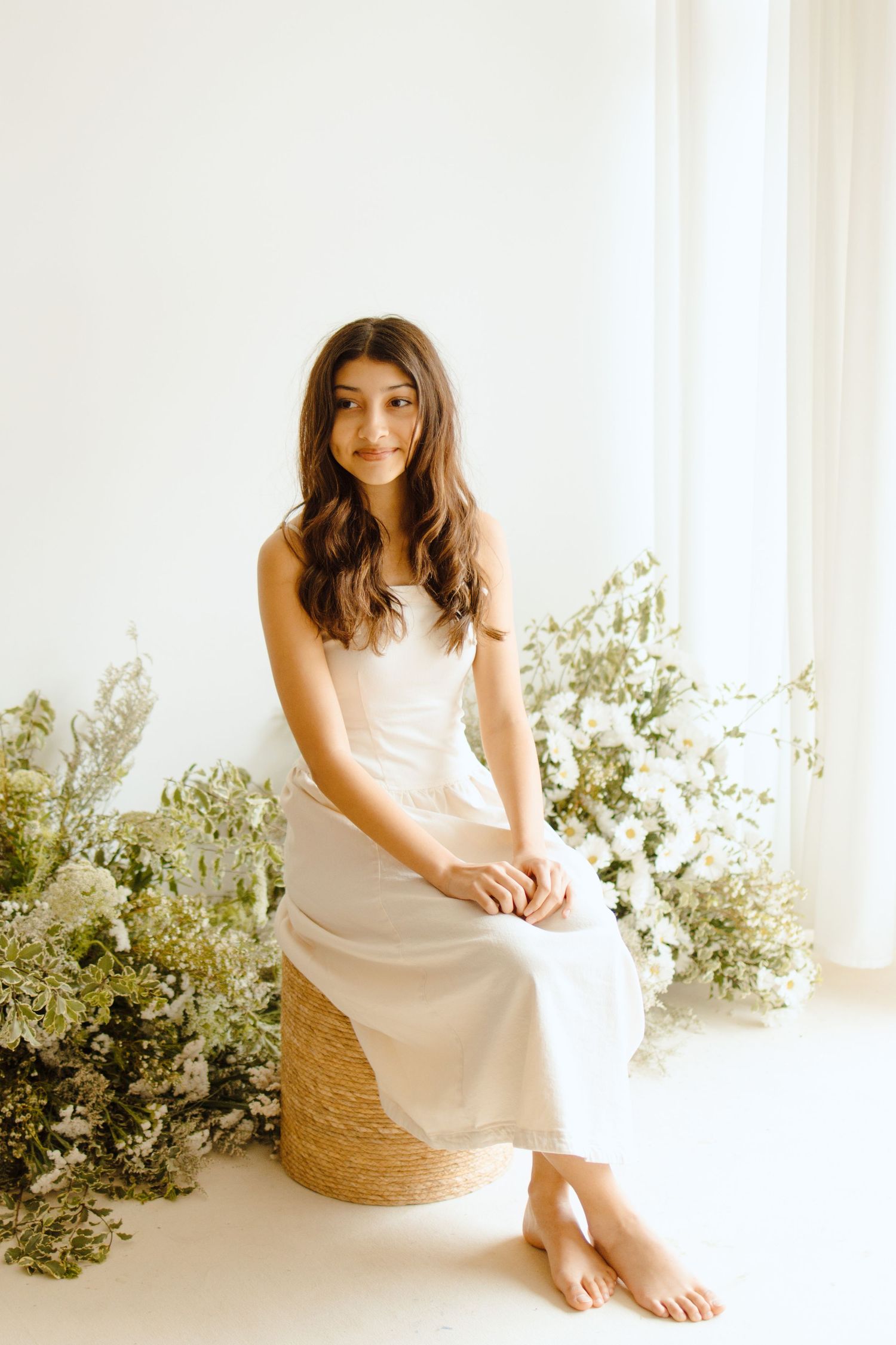 Young woman in white slip dress seated on wicker stool surrounded by white florals in bright studio.