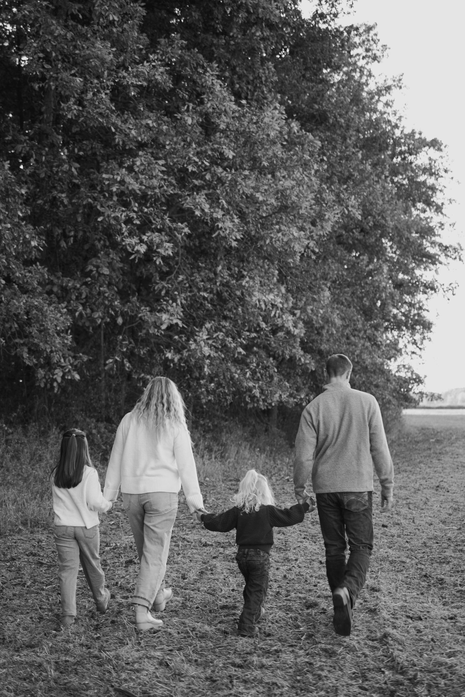 Black and white photo of a family of four walking hand-in-hand through a field toward trees.