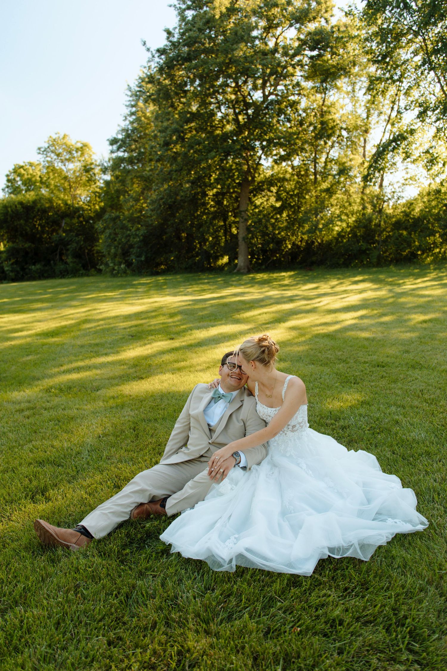 Bride and groom sitting together on lush green grass outdoors, bride in white gown, groom in tan suit with bow tie.