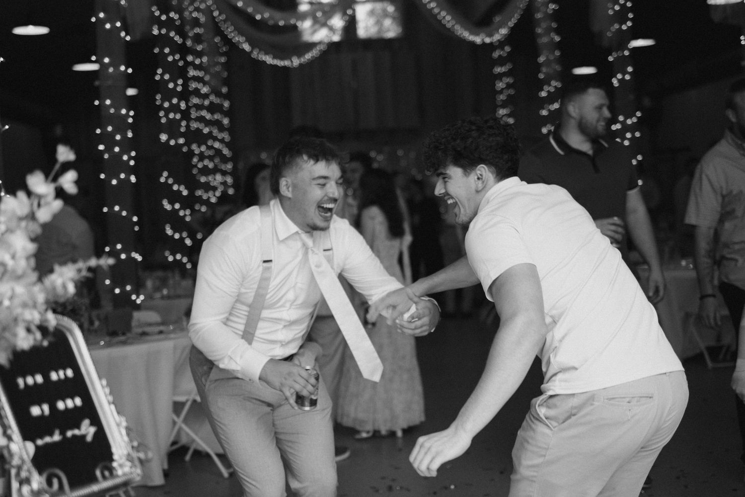 Two men dancing joyfully at wedding reception with string lights in background.