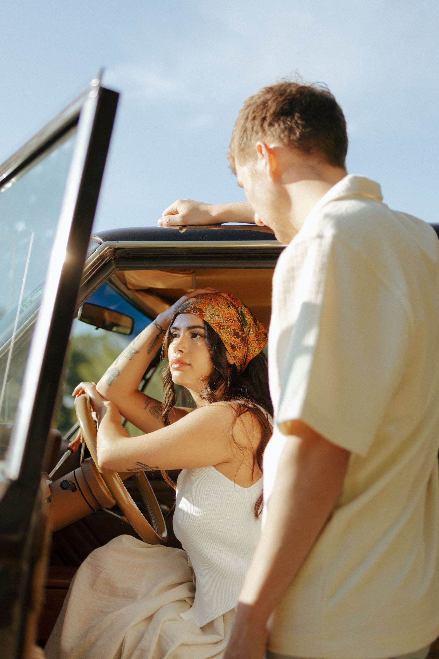 A woman in a white dress and orange headscarf sits in a vintage car, looking up at a man standing by the open door.