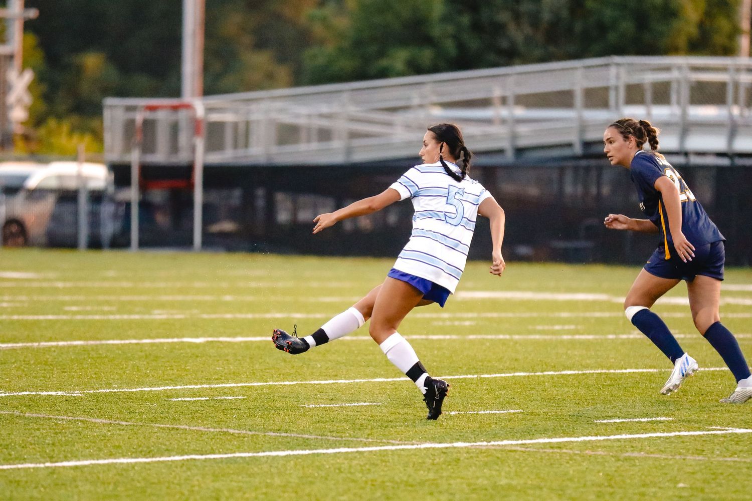 Soccer player in white uniform kicks ball on field while opponent in blue jersey defends during match.
