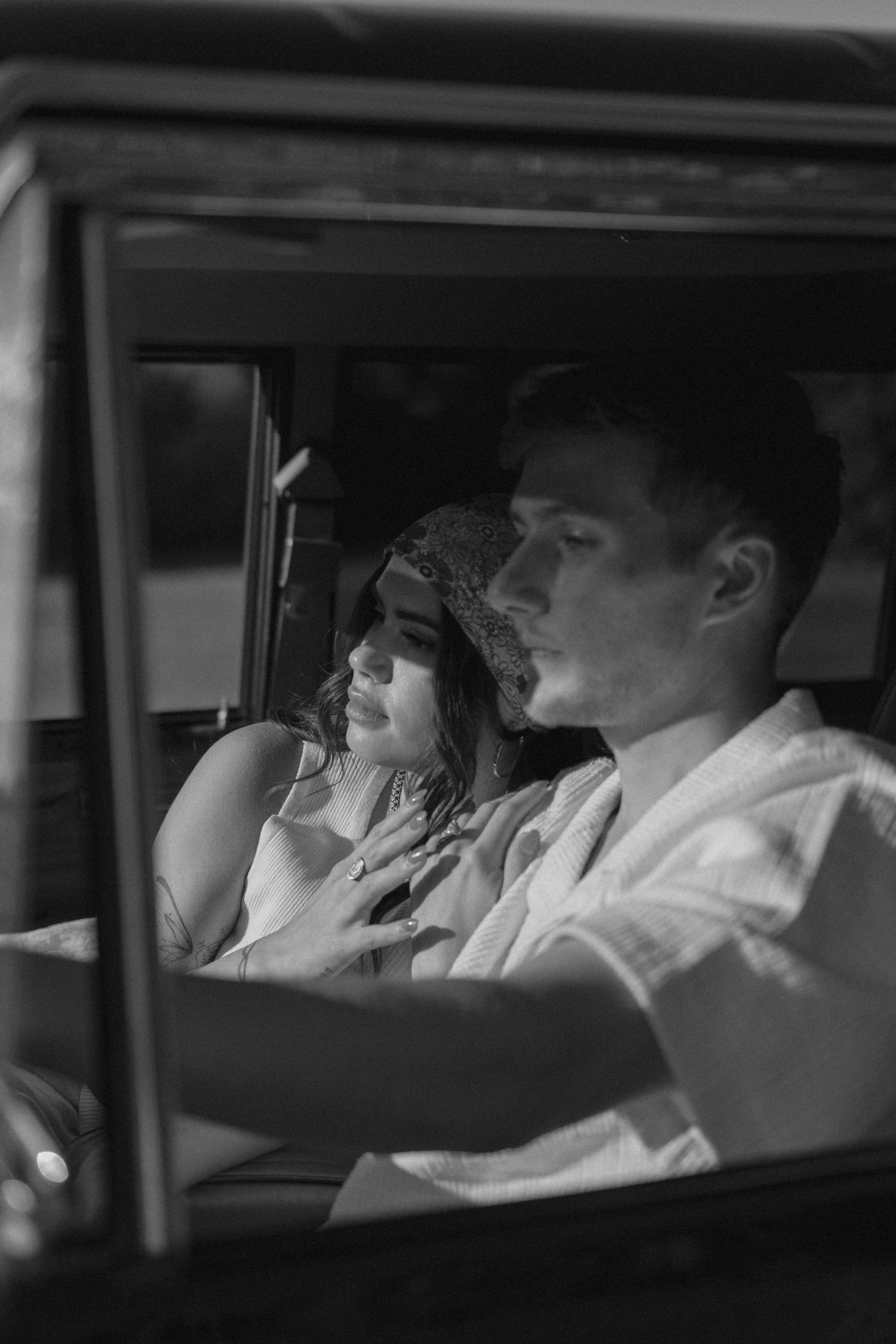 Black and white photo of a couple sitting together in a vintage car, woman in hat leaning toward man.
