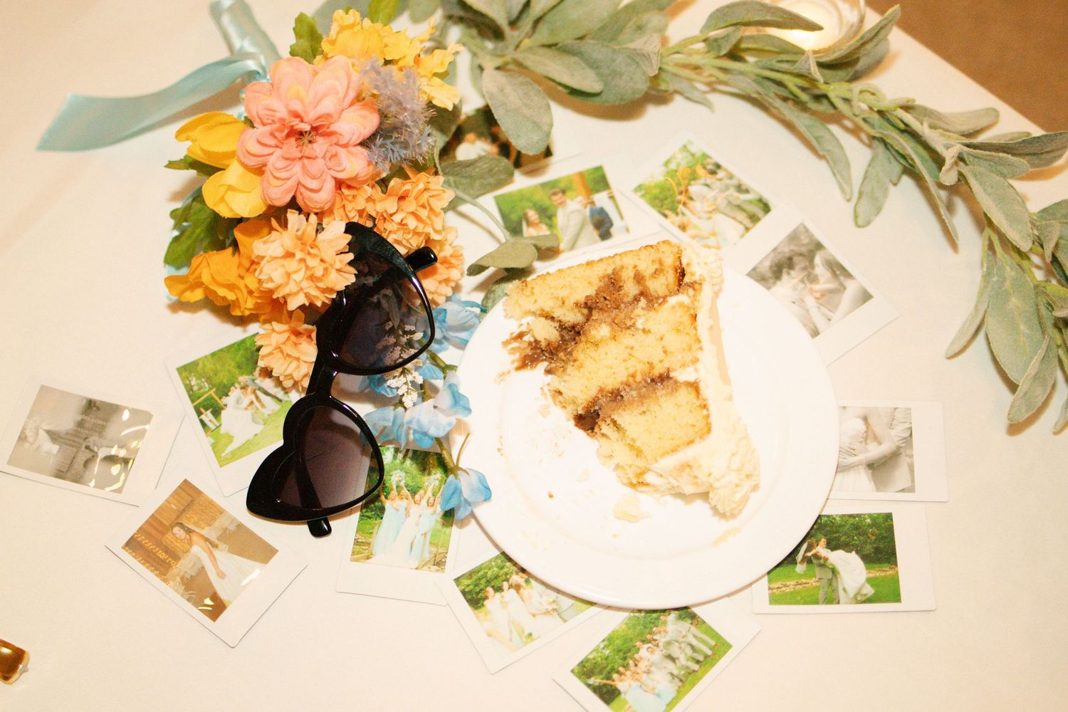 Wedding reception table detail with cake slice, sunglasses, floral arrangement, and scattered Polaroid photos.