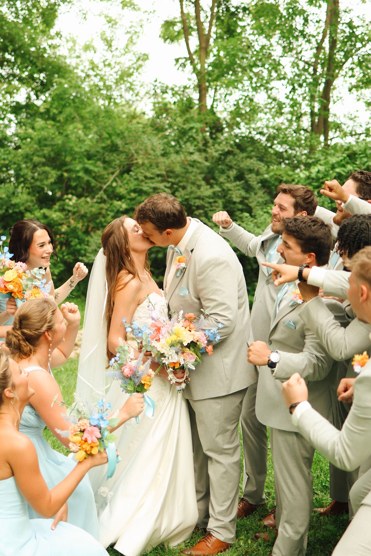 A lively wedding party celebrates outdoors with pastel bridesmaid dresses and colorful bouquets against a green forest backdrop.