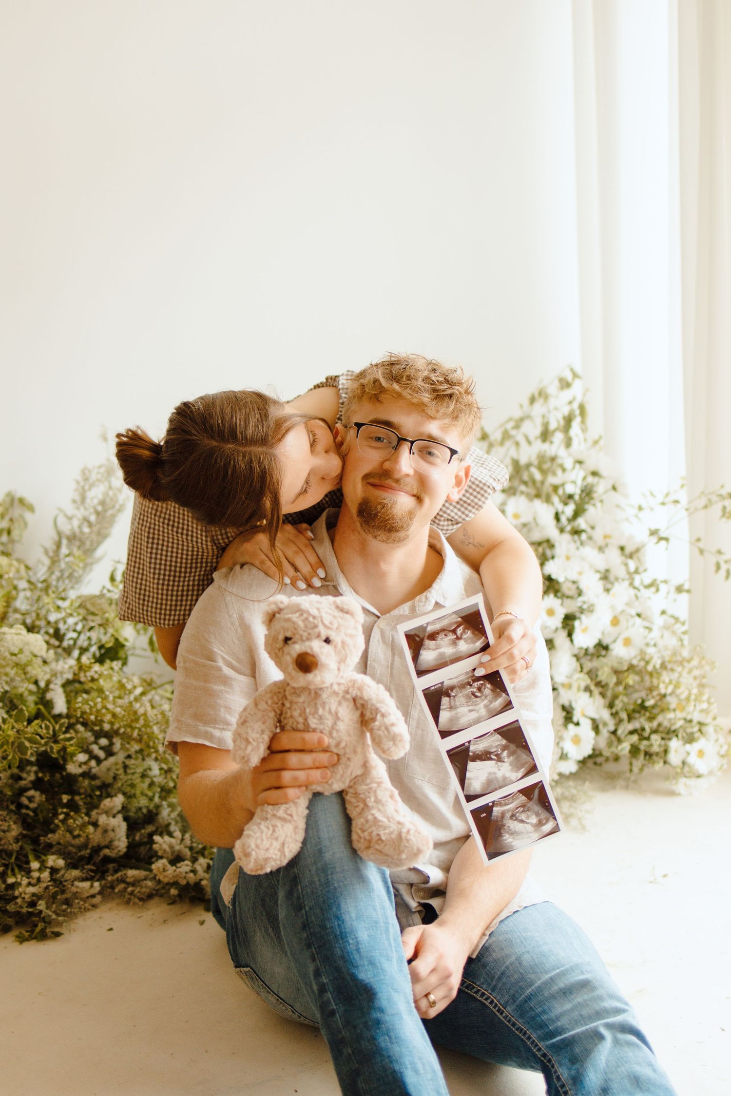 Couple sitting on floor with ultrasound photos and teddy bear, surrounded by flowers, announcing pregnancy.
