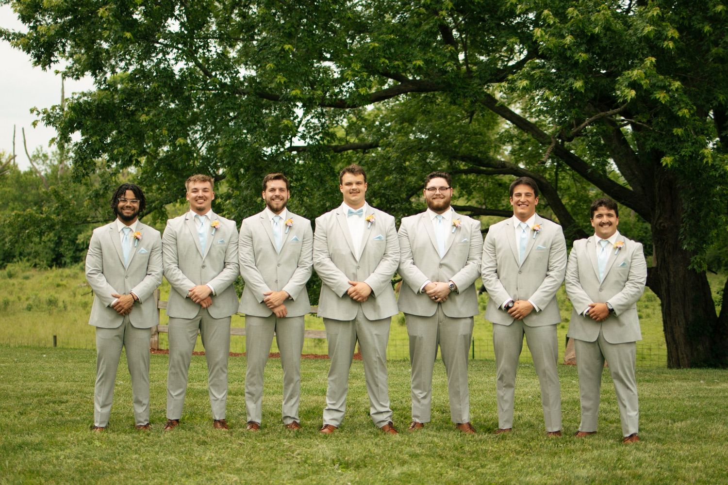Groom and six groomsmen in matching light gray suits pose together outdoors under large green tree.