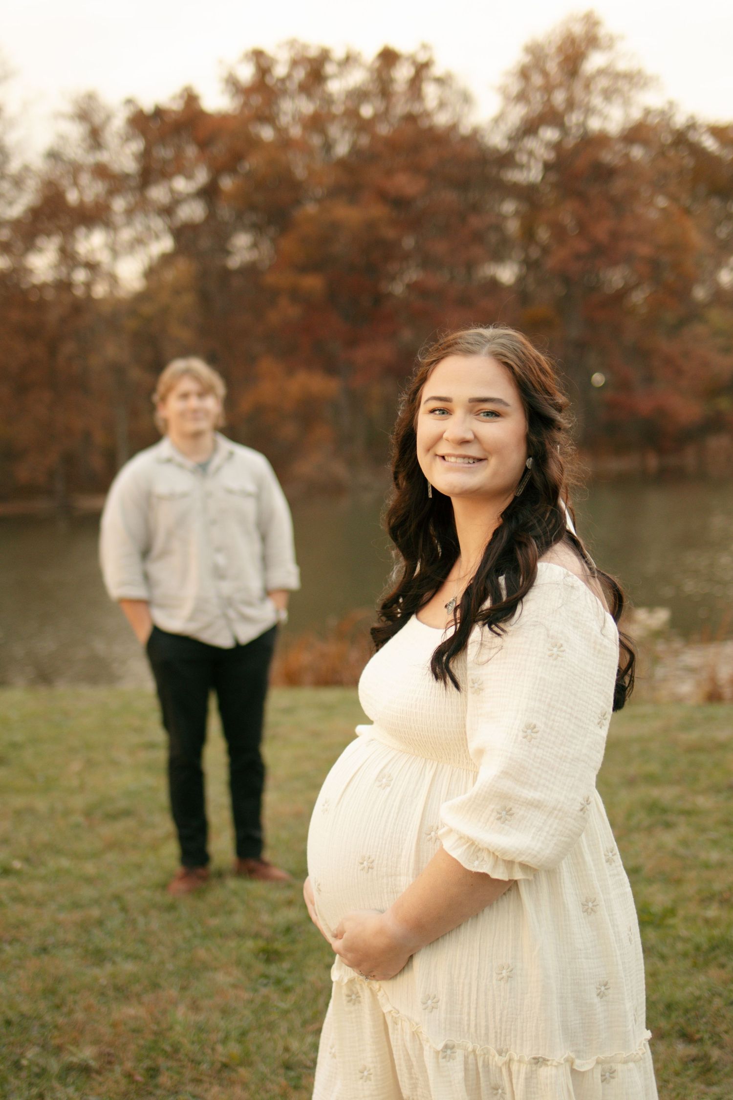 Pregnant woman in white dress smiling outdoors in autumn park with man standing near lake in background.