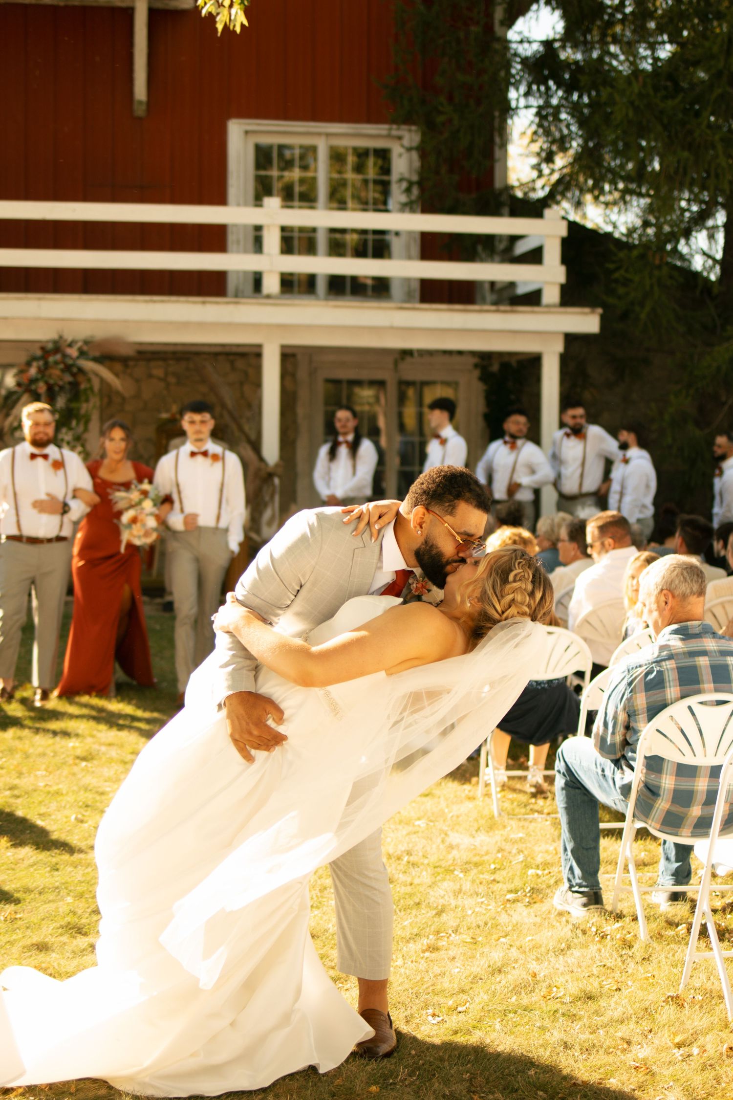 Groom dips bride for a kiss during outdoor wedding ceremony at rustic red barn venue with wedding party watching.