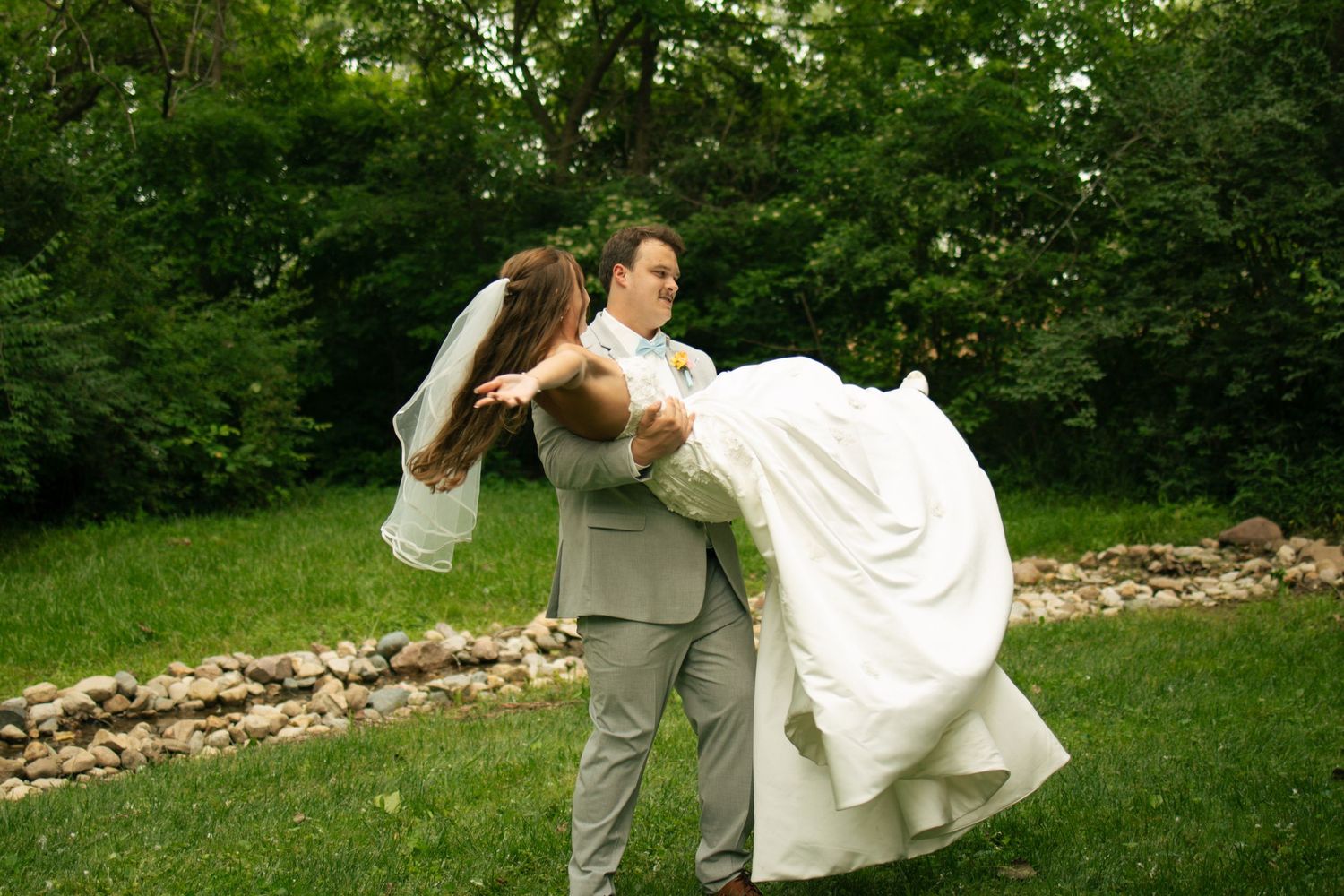 Groom in gray suit sweeping bride in white wedding dress off her feet outdoors in lush green garden setting.