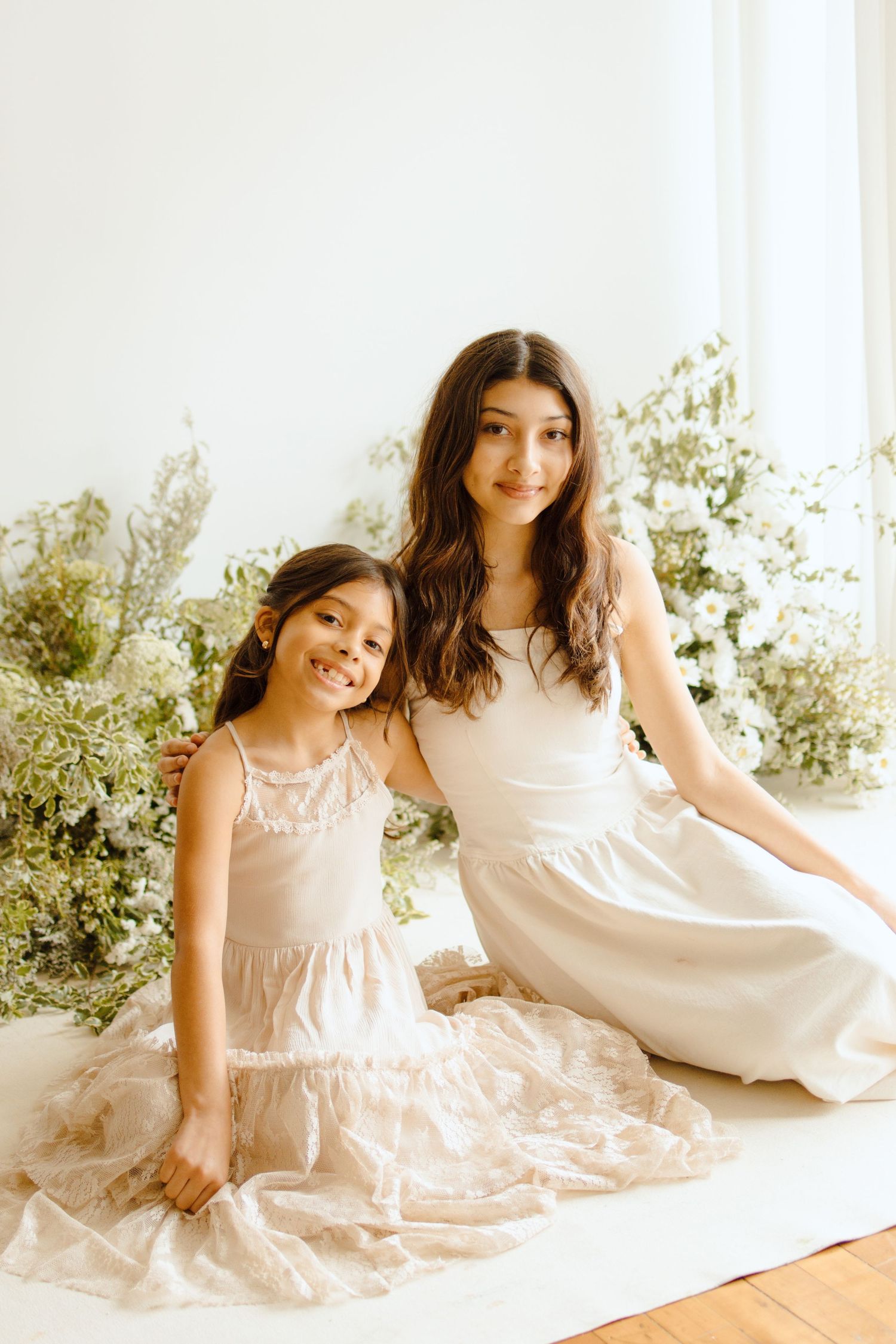 Two young girls in white and blush dresses pose together among white flowers in a bright studio setting.
