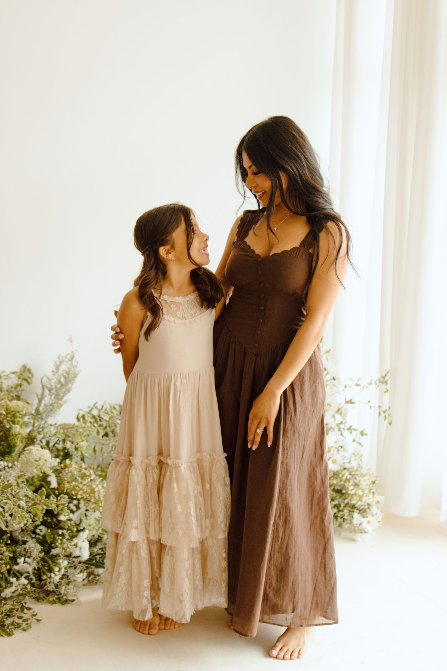 Mother and daughter in matching boho dresses, smiling together surrounded by white flowers in a bright studio.