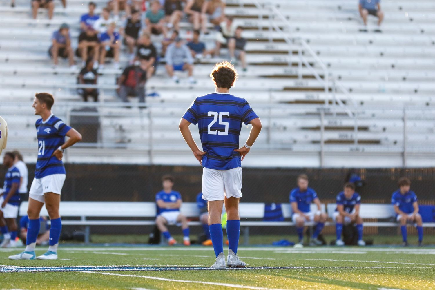 Soccer player wearing jersey number 25 stands alone on field while team sits on sideline bench in background.