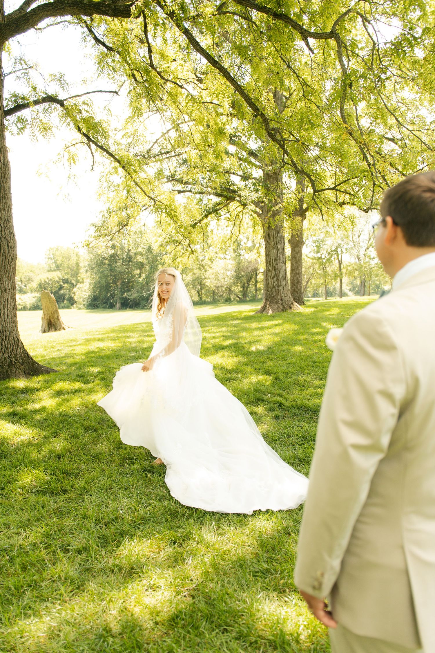 Bride in white gown and veil walks toward groom in tan suit under lush green trees on sunny wedding day.