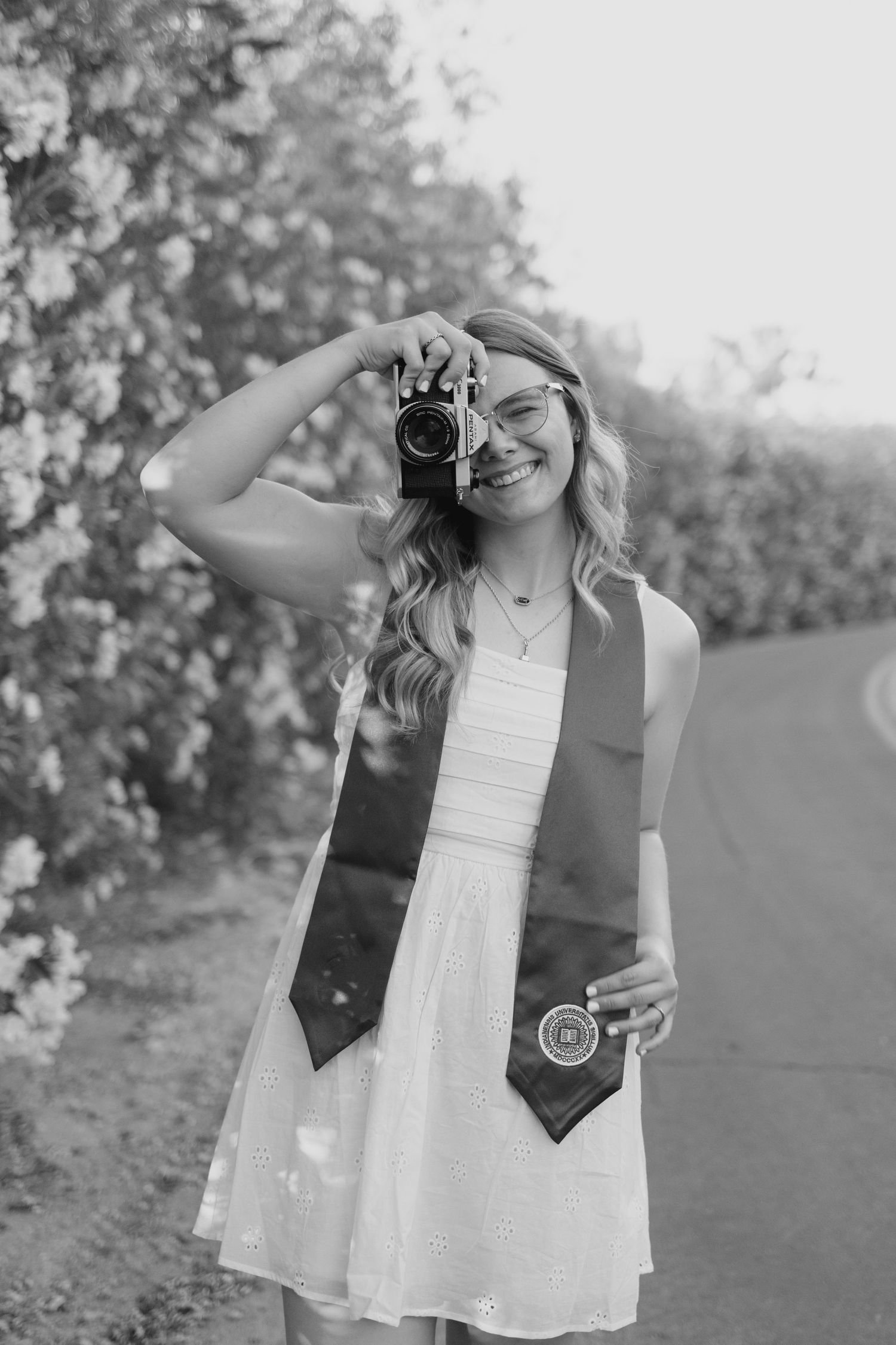 A person in a white dress and vest holds up a vintage camera while standing in a garden setting.