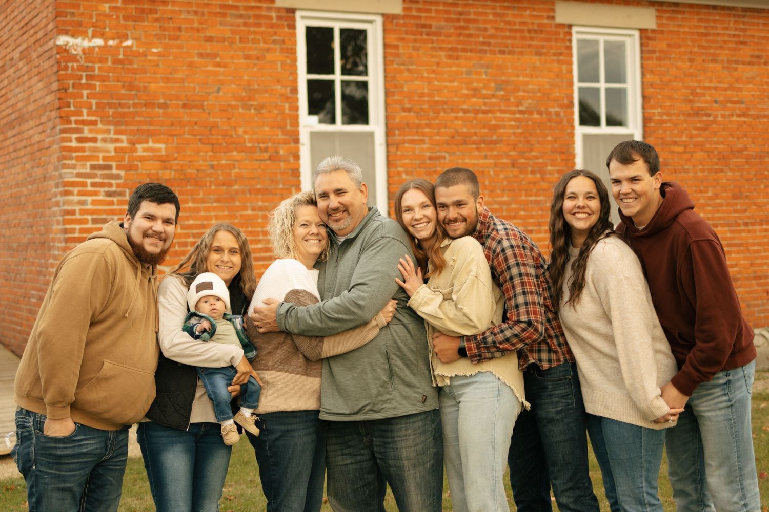 Large family group smiling together outdoors in front of a red brick building in fall.