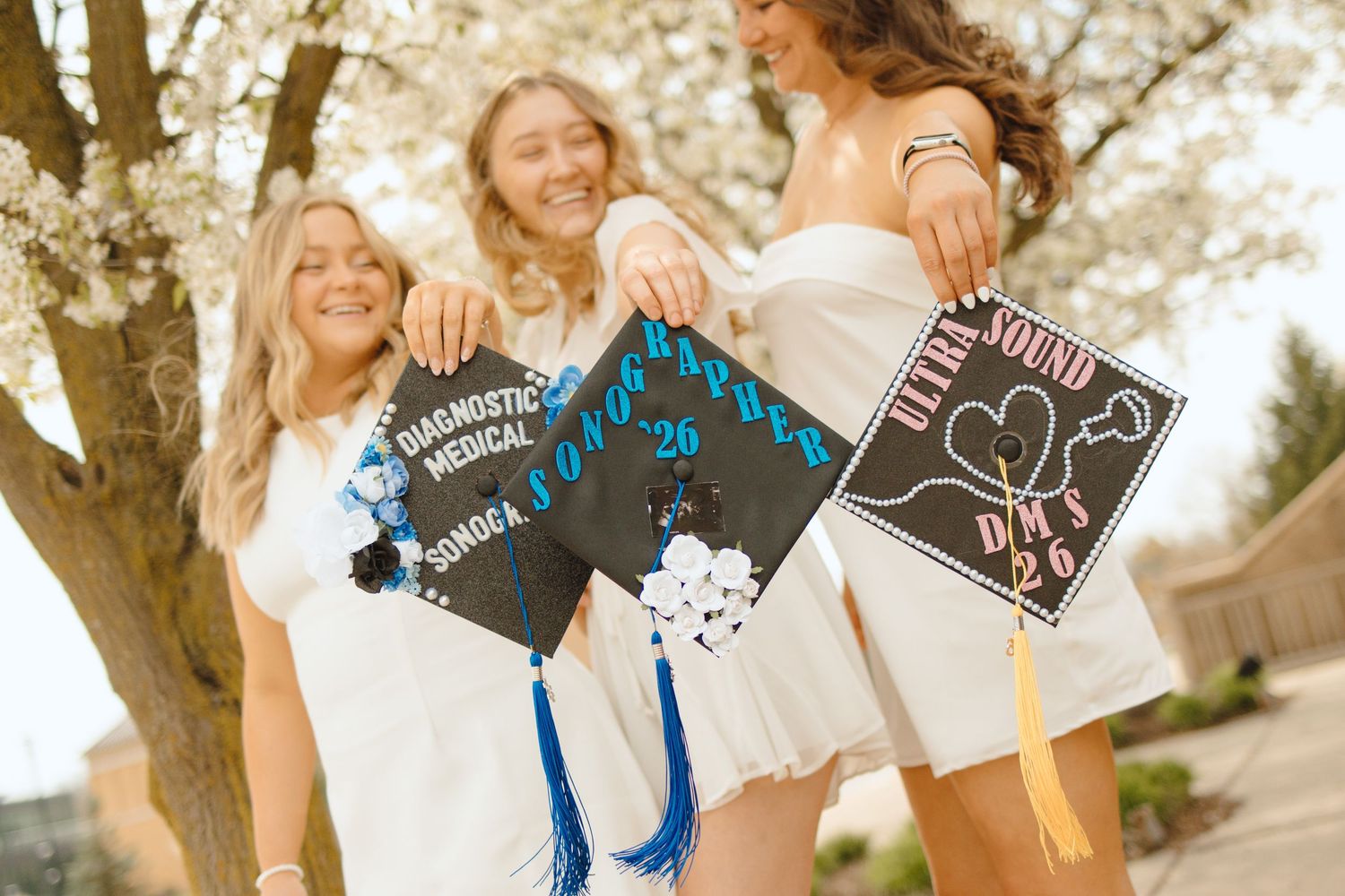 Three smiling graduates in white dresses proudly display decorated black mortarboard caps with blue tassels outdoors.