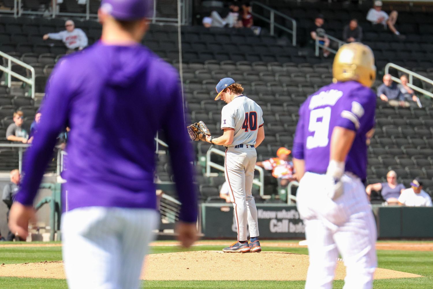 Baseball players in purple uniforms and a player in white uniform stand on a field with stadium seating in the background.
