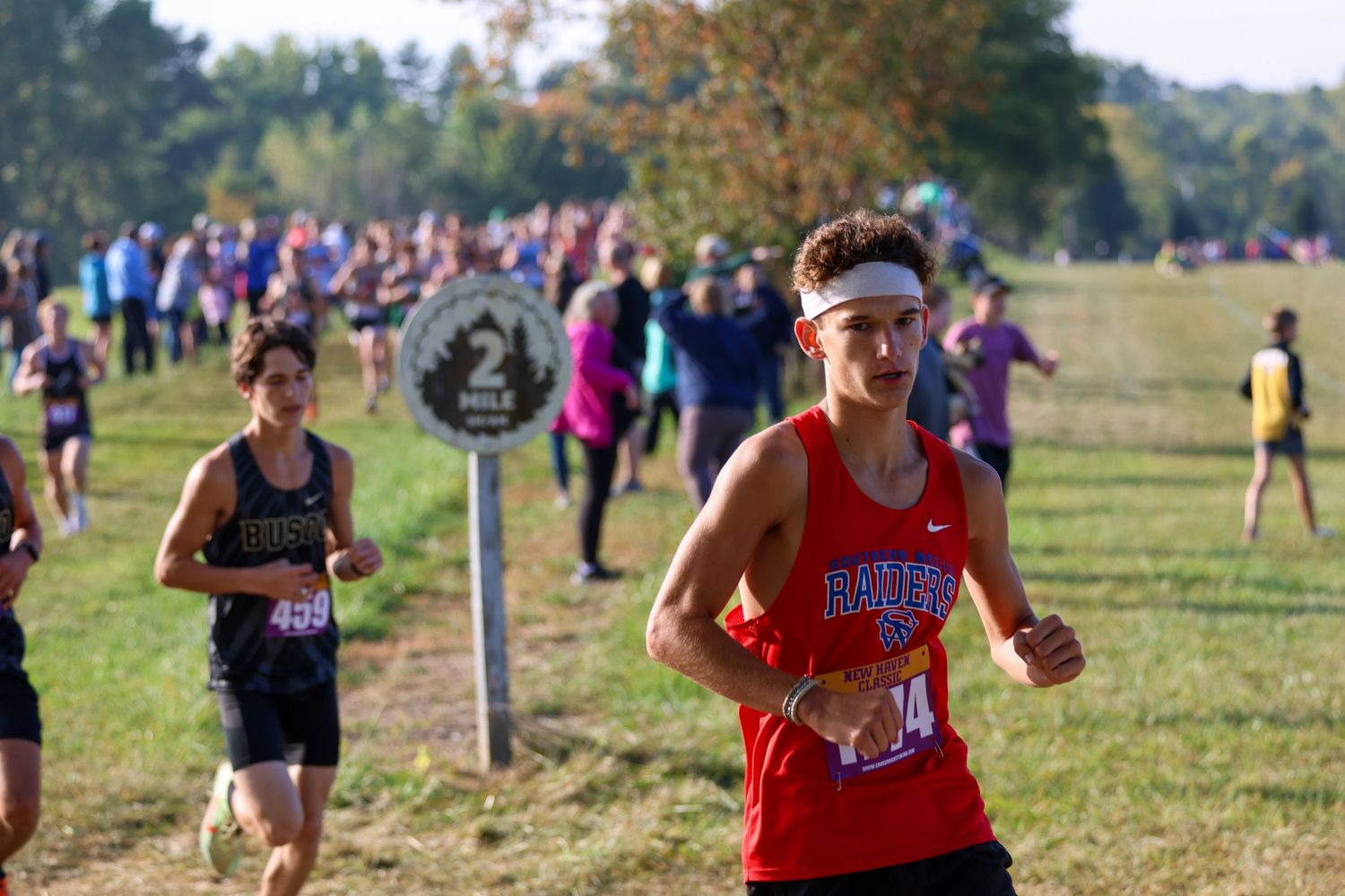Cross country runners competing on a grassy course during a sunny fall day with spectators visible in background.