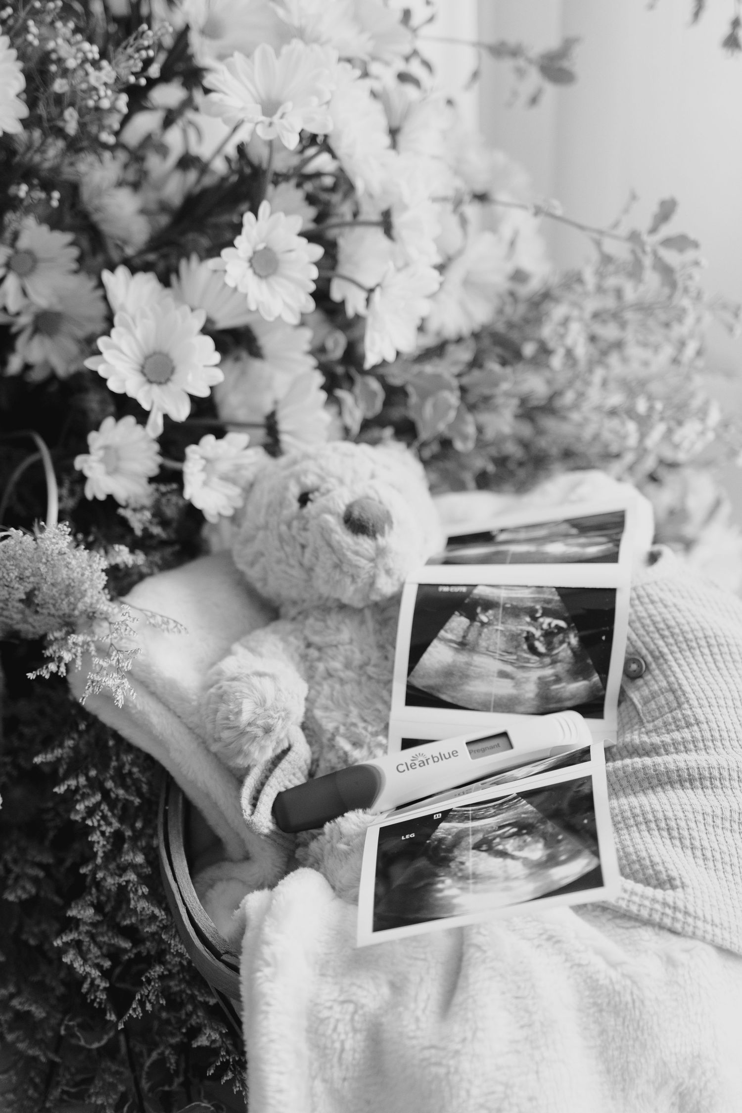 Black and white photo of ultrasound scans, teddy bear, pregnancy test, and flowers arranged together.