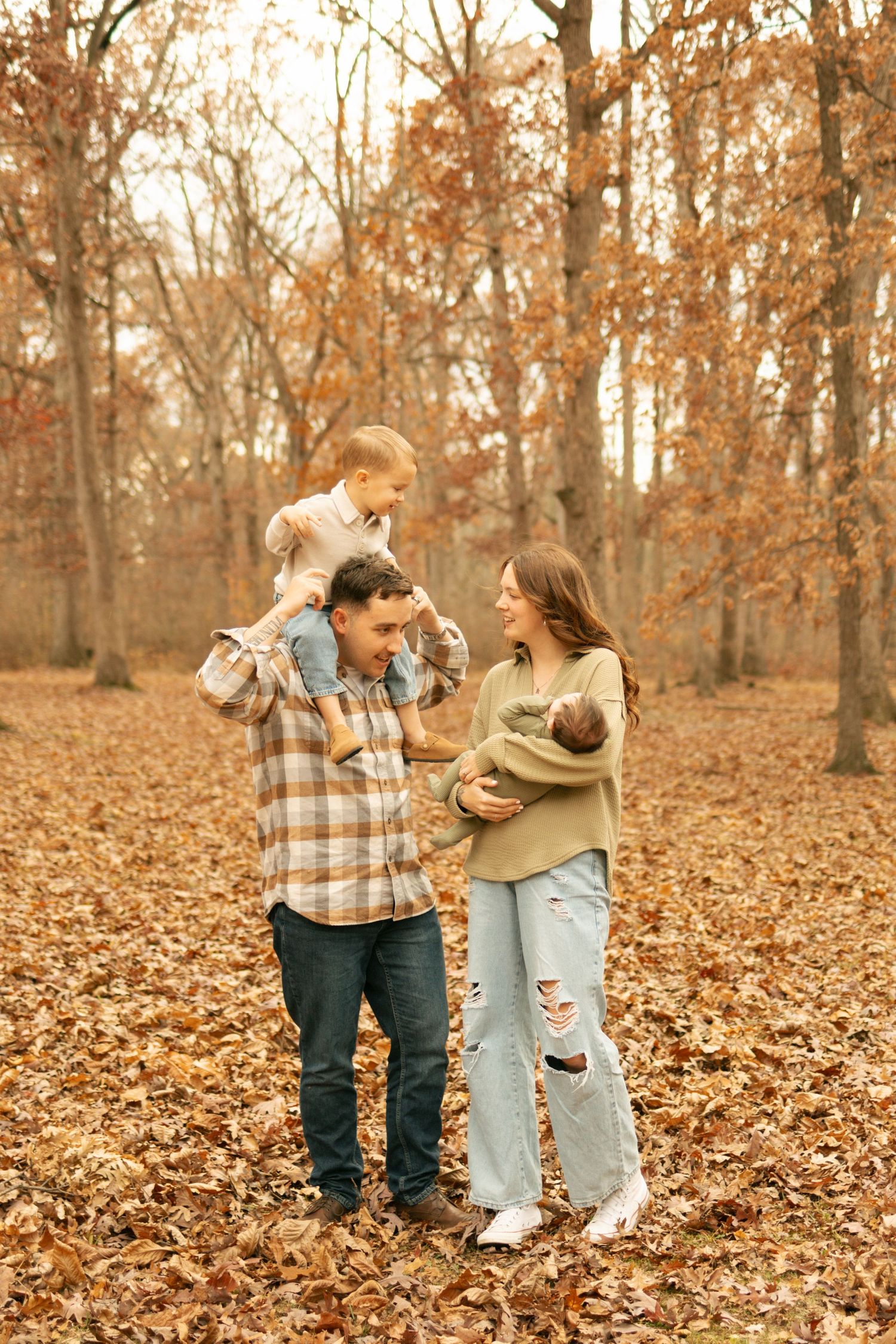 Family of four in autumn forest, dad carrying toddler on shoulders while mom holds newborn baby.
