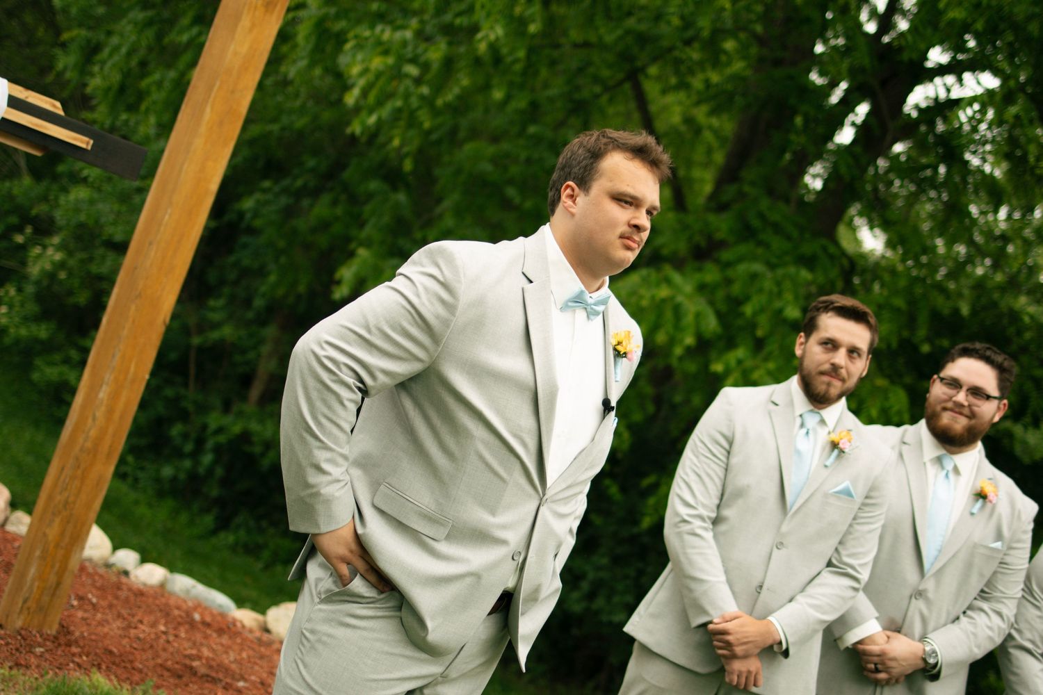 Groomsmen in matching light gray suits with bow ties and boutonnieres standing outdoors at a wedding ceremony.