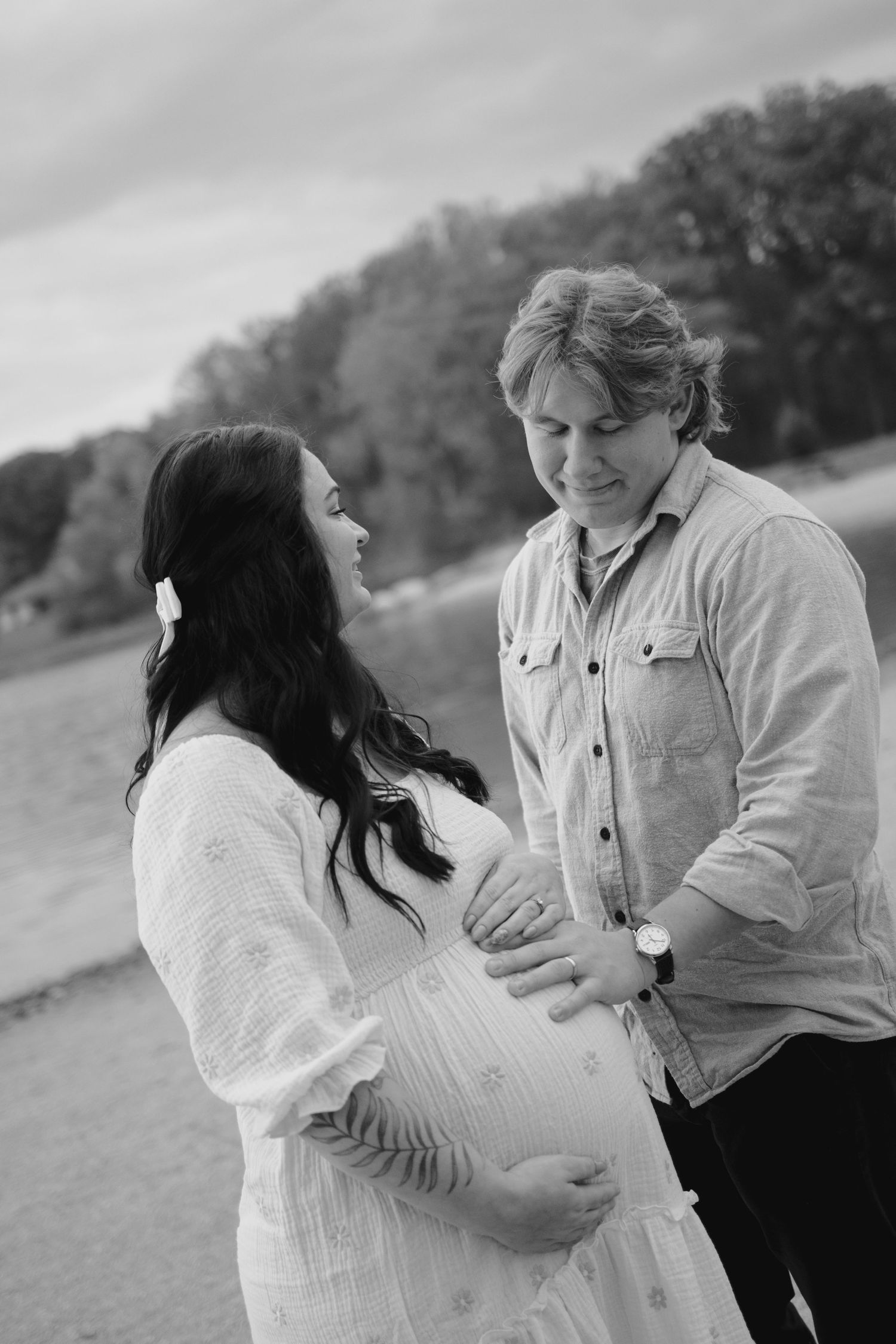 Black and white maternity photo of a couple smiling at each other, hands on pregnant belly, outdoors near water.