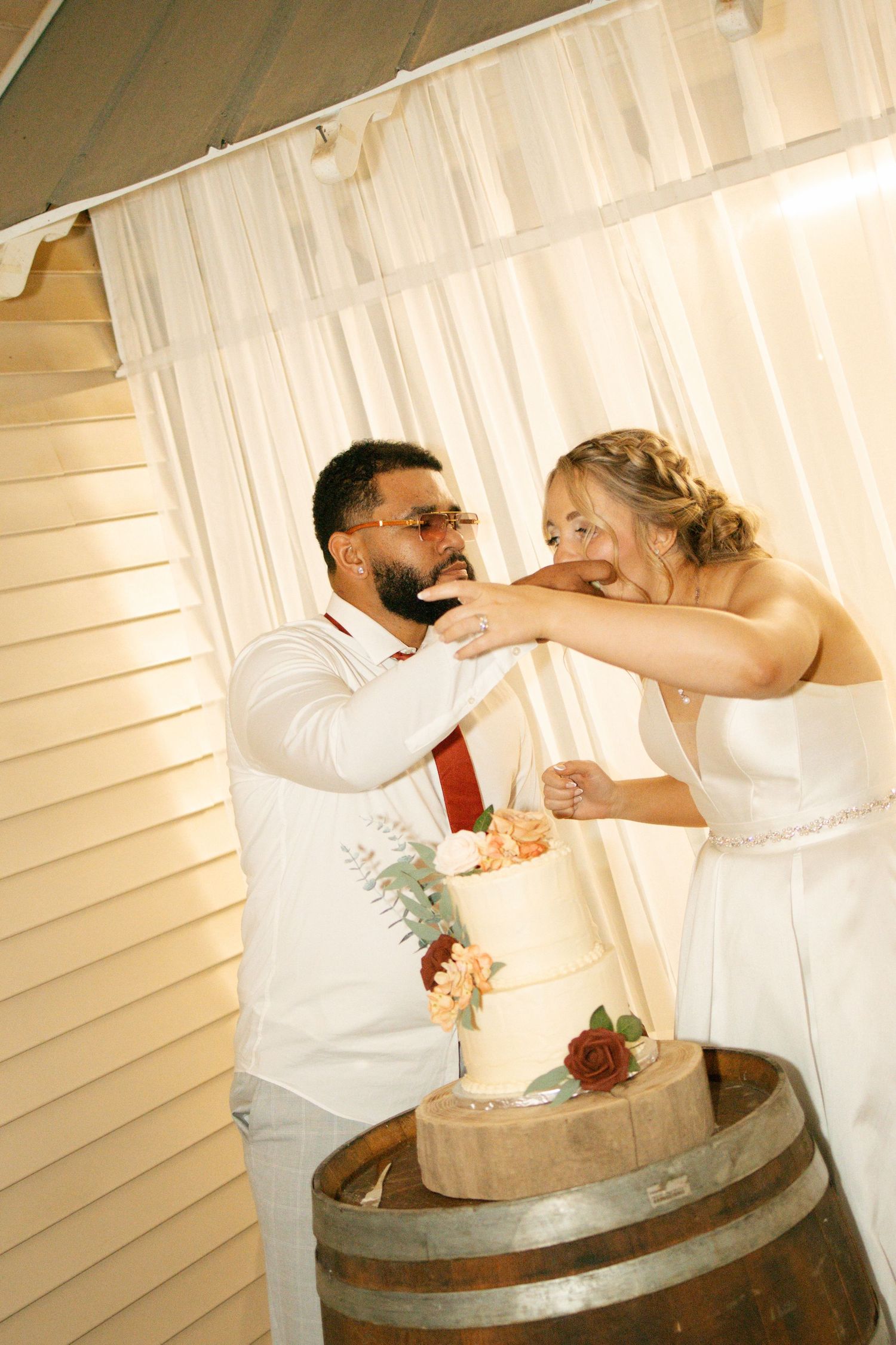 Bride and groom playfully feed each other wedding cake atop a rustic barrel at their reception.
