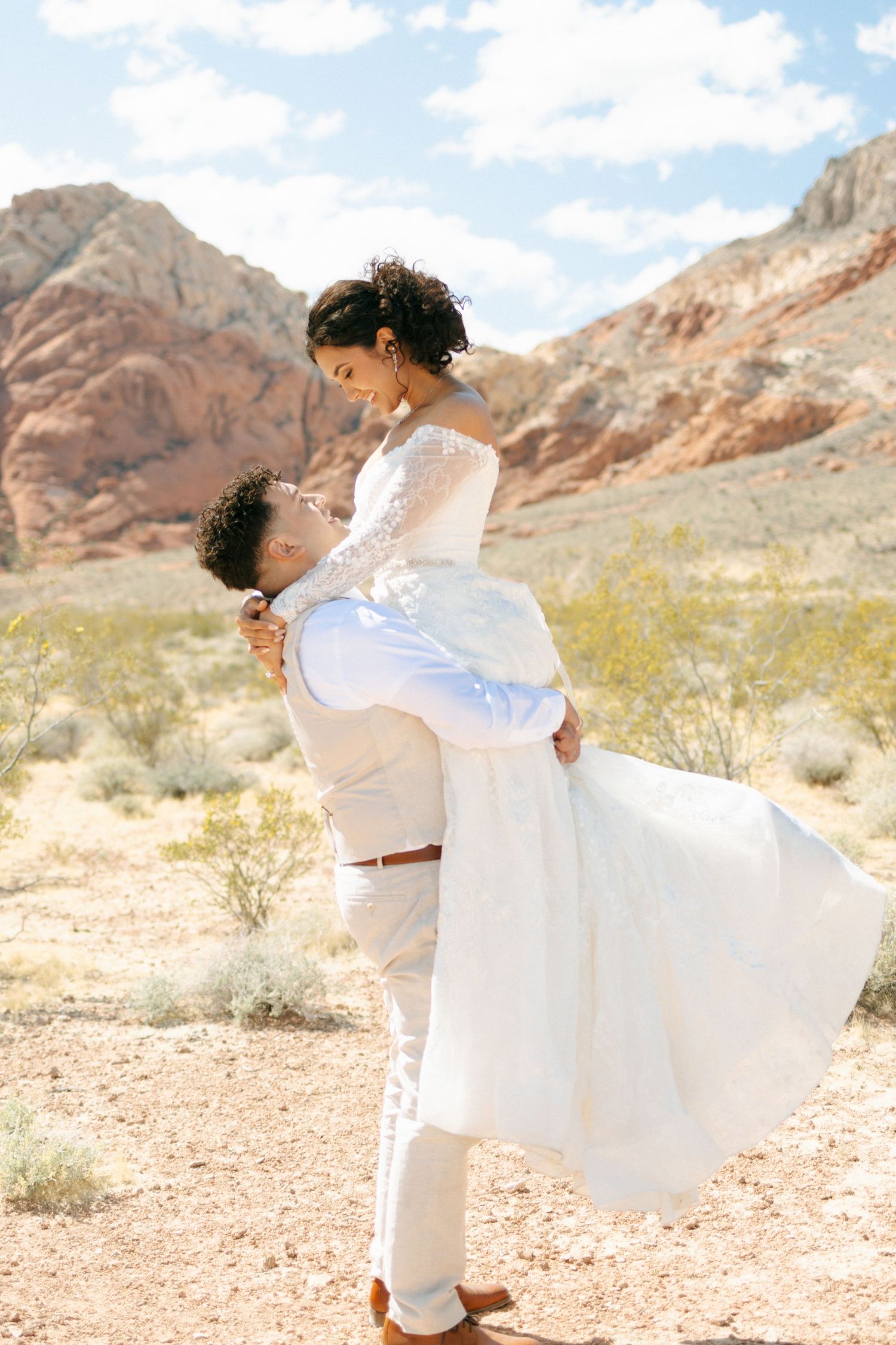 Newlyweds embrace in a romantic moment against red rock mountains and desert landscape at sunset.