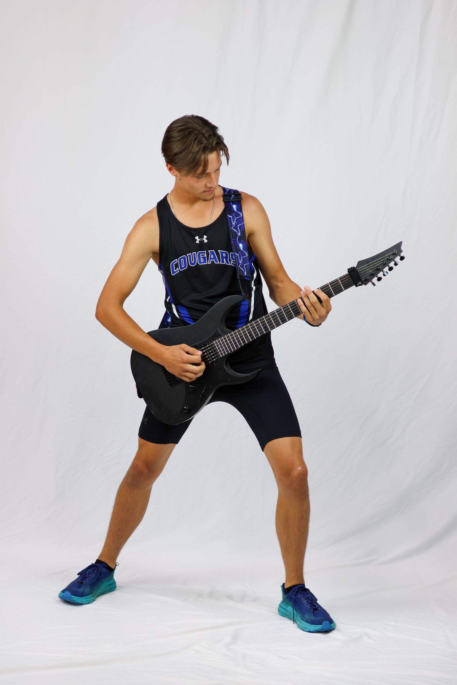 Guitarist in athletic wear plays a black electric guitar against a white backdrop while striking a dynamic pose.