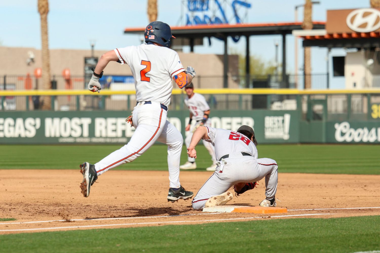 Baseball game action shows player running to base as opposing player attempts a slide during fielding play.