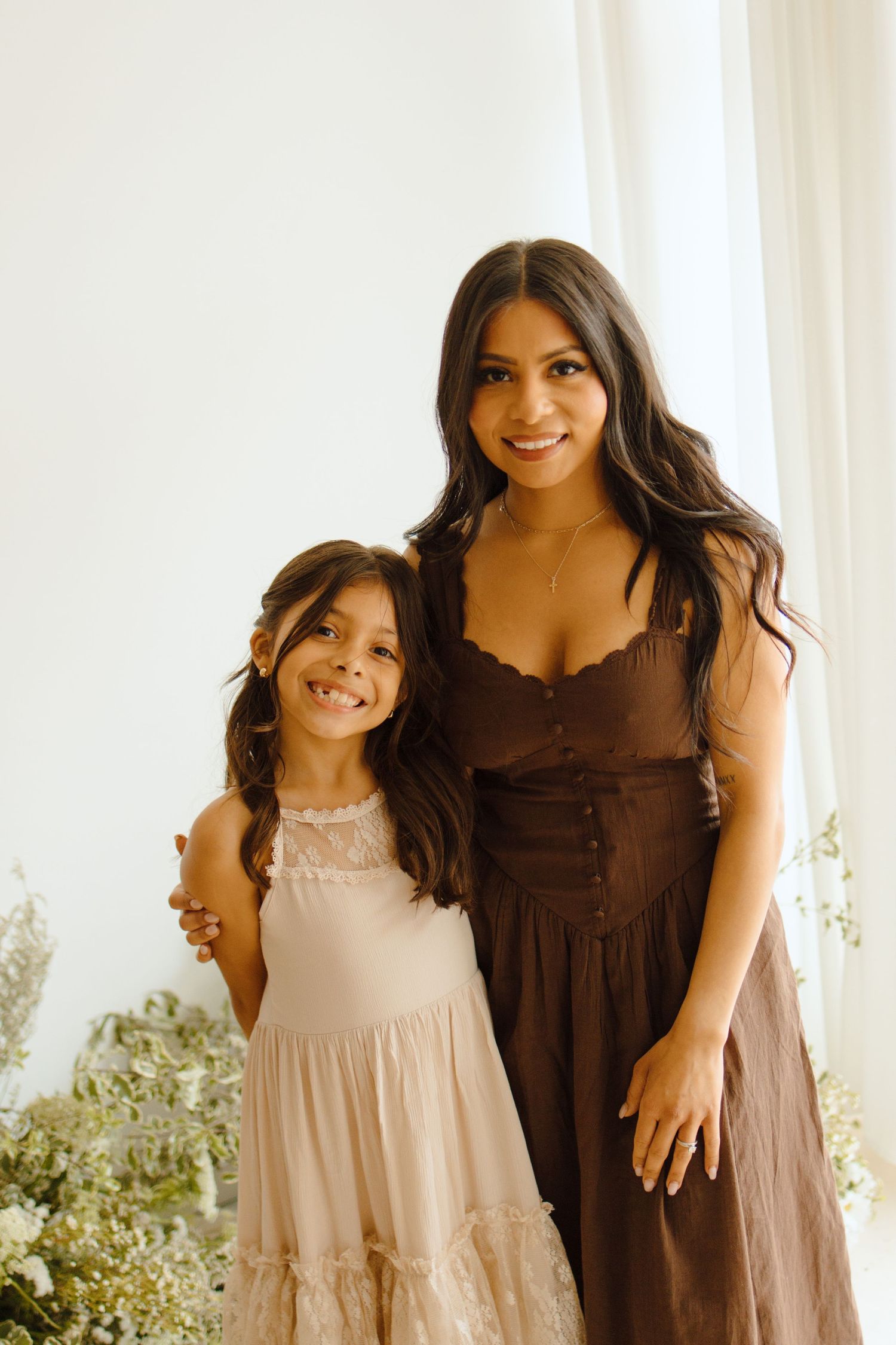 Woman in brown dress and young girl in pink dress smiling together in bright studio setting.