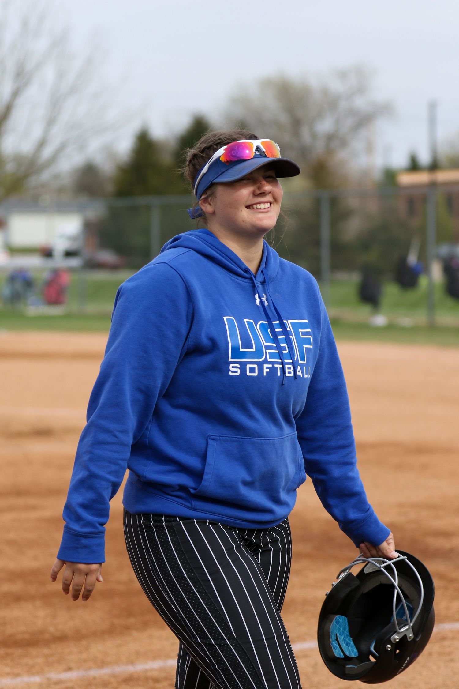 Softball player in blue LIFE hoodie and pinstriped pants smiles on dirt field during practice or game.