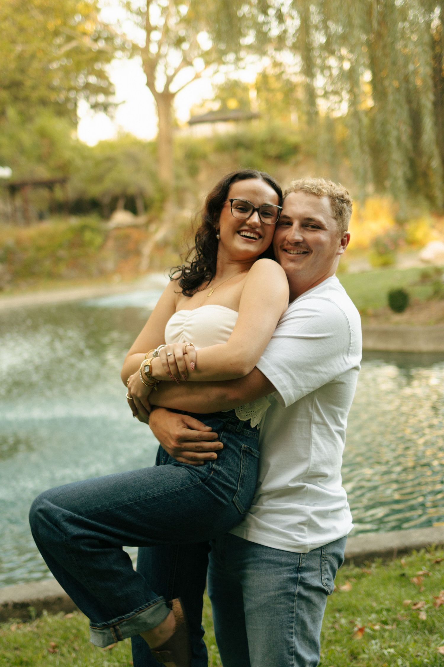 A happy couple embraces and poses together in front of a serene pond in a park.