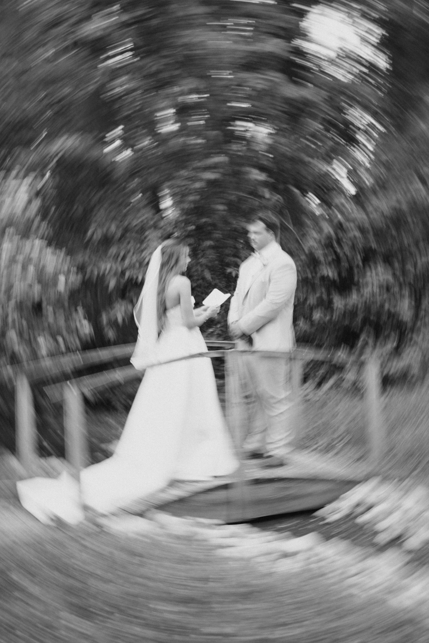 Black and white swirling artistic photo of bride and groom exchanging vows on a wooden bridge outdoors.