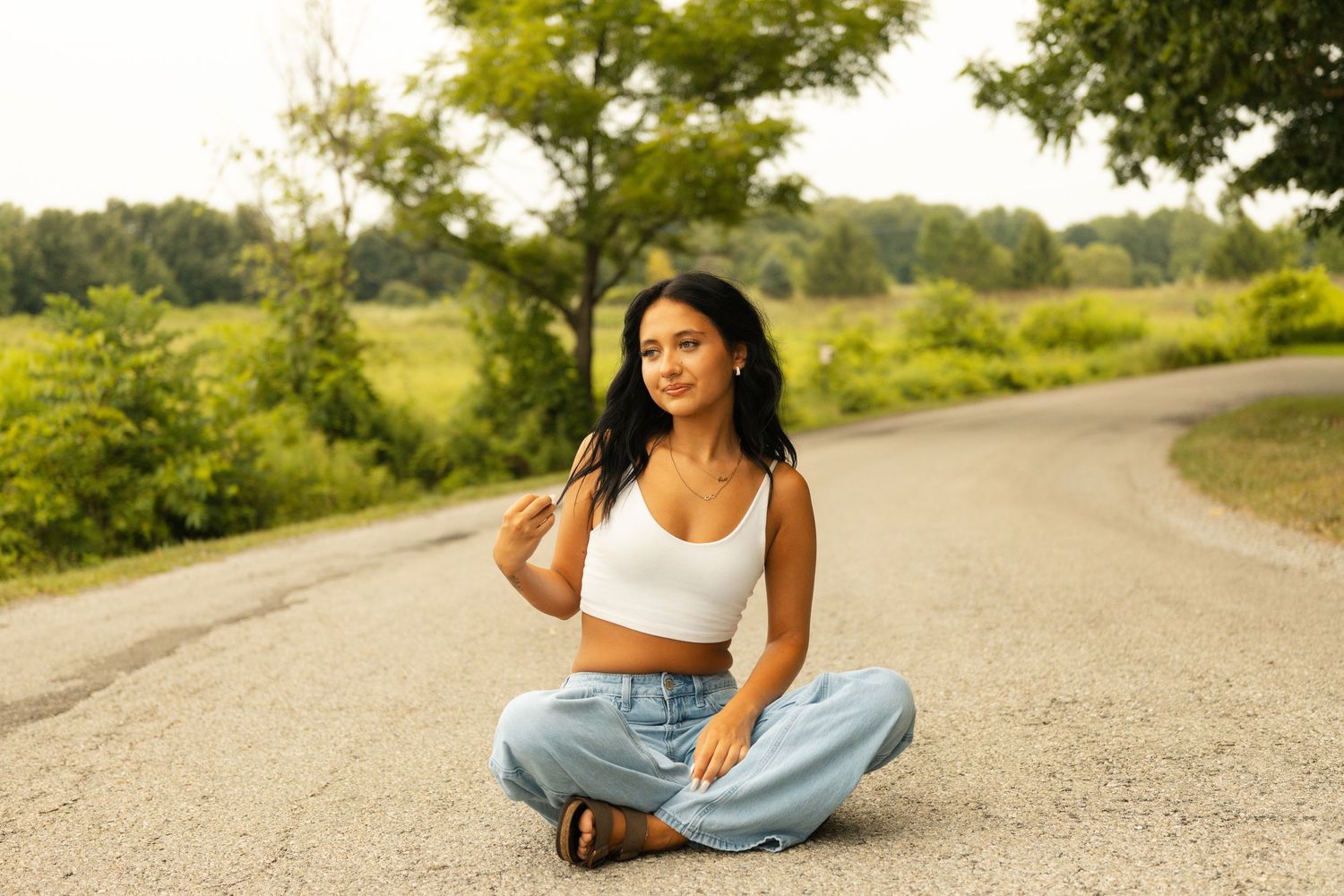 Young woman in white crop top and jeans sitting cross-legged on a road surrounded by lush green trees.